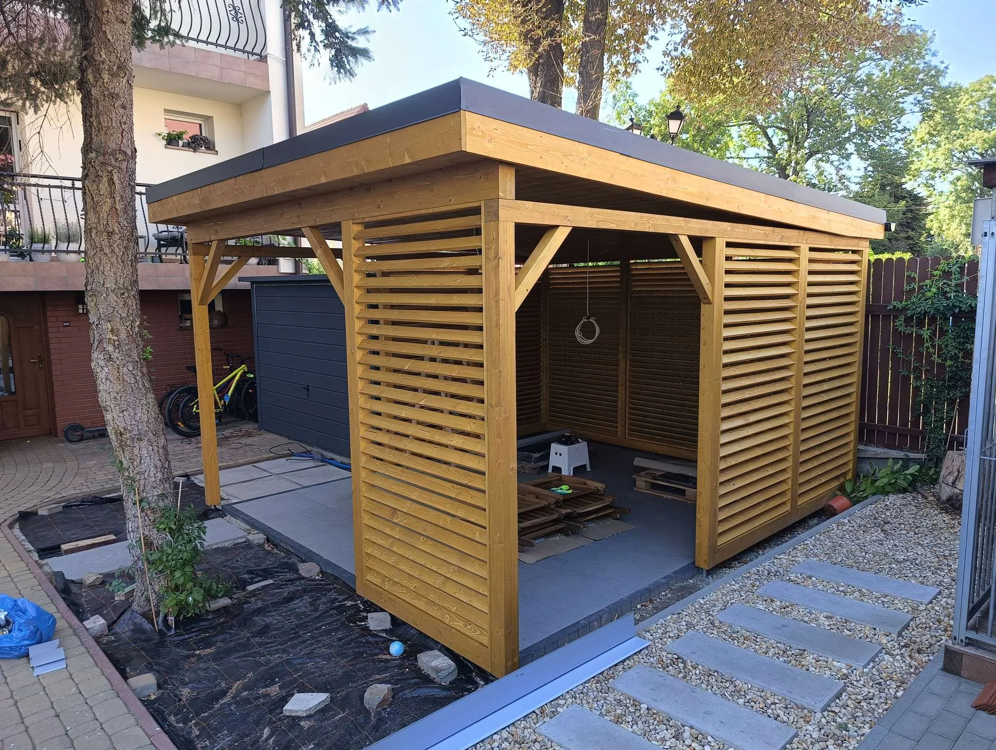 A wooden structure under construction in a backyard with trees and a neighboring building in the background. The structure has a flat roof with black edging and slatted side walls.