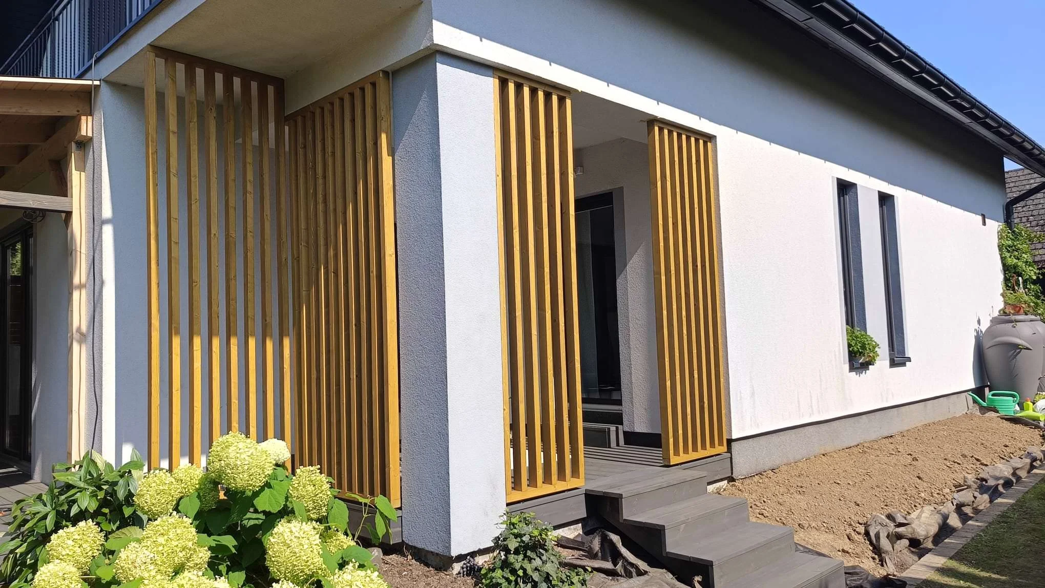 Modern house exterior with white walls, black trim, and wooden slats on the patio, surrounded by plants and gardening tools.