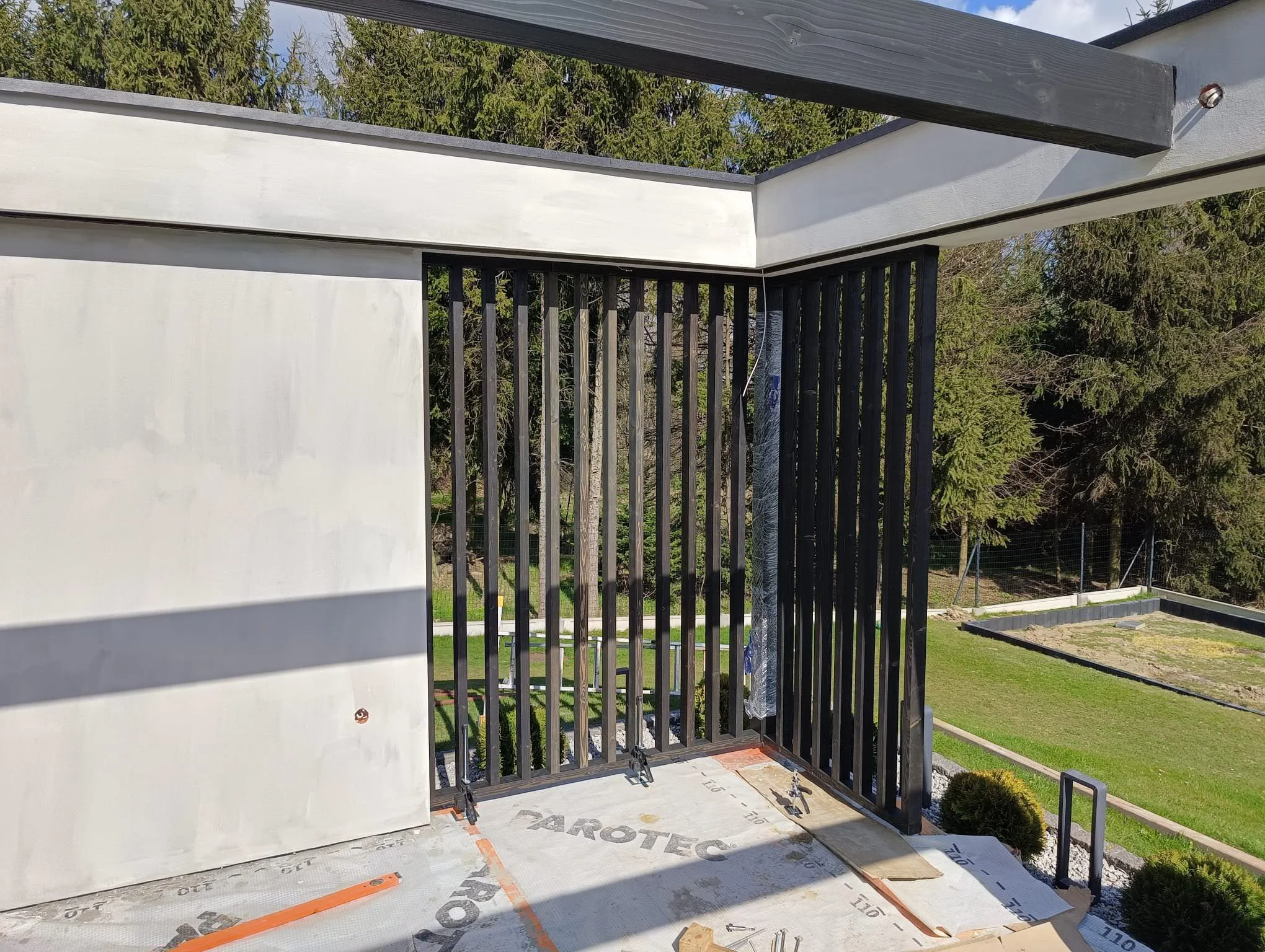 Photo showing a balcony under construction with black metal railing, white wall, and partially installed flooring, with trees and blue sky in the background.
