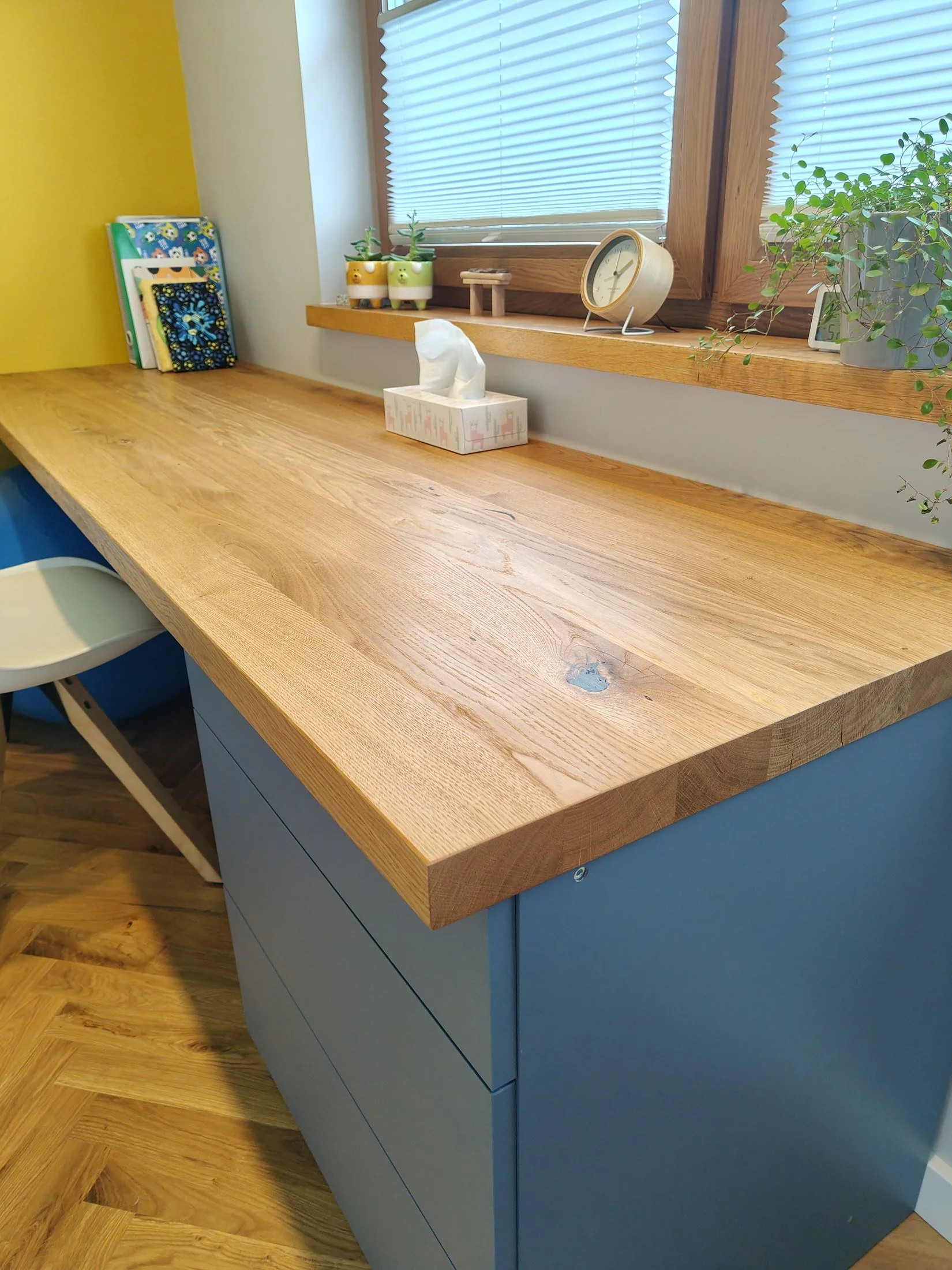 A wooden countertop with a tissue box, small potted plants, and a clock next to a window with blinds. The wall behind is painted yellow, and there is a blue cabinet below the countertop. The floor is wooden.