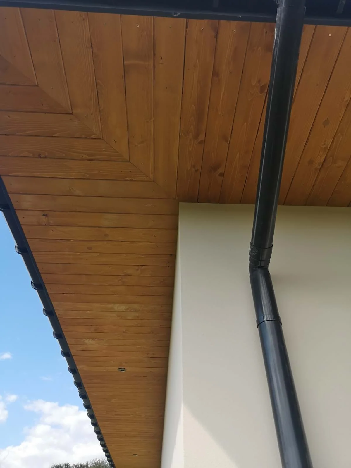 A view of a house's exterior showing a wooden ceiling with a vented overhang and a black drainpipe attached to the wall, with partly cloudy blue sky in the background.