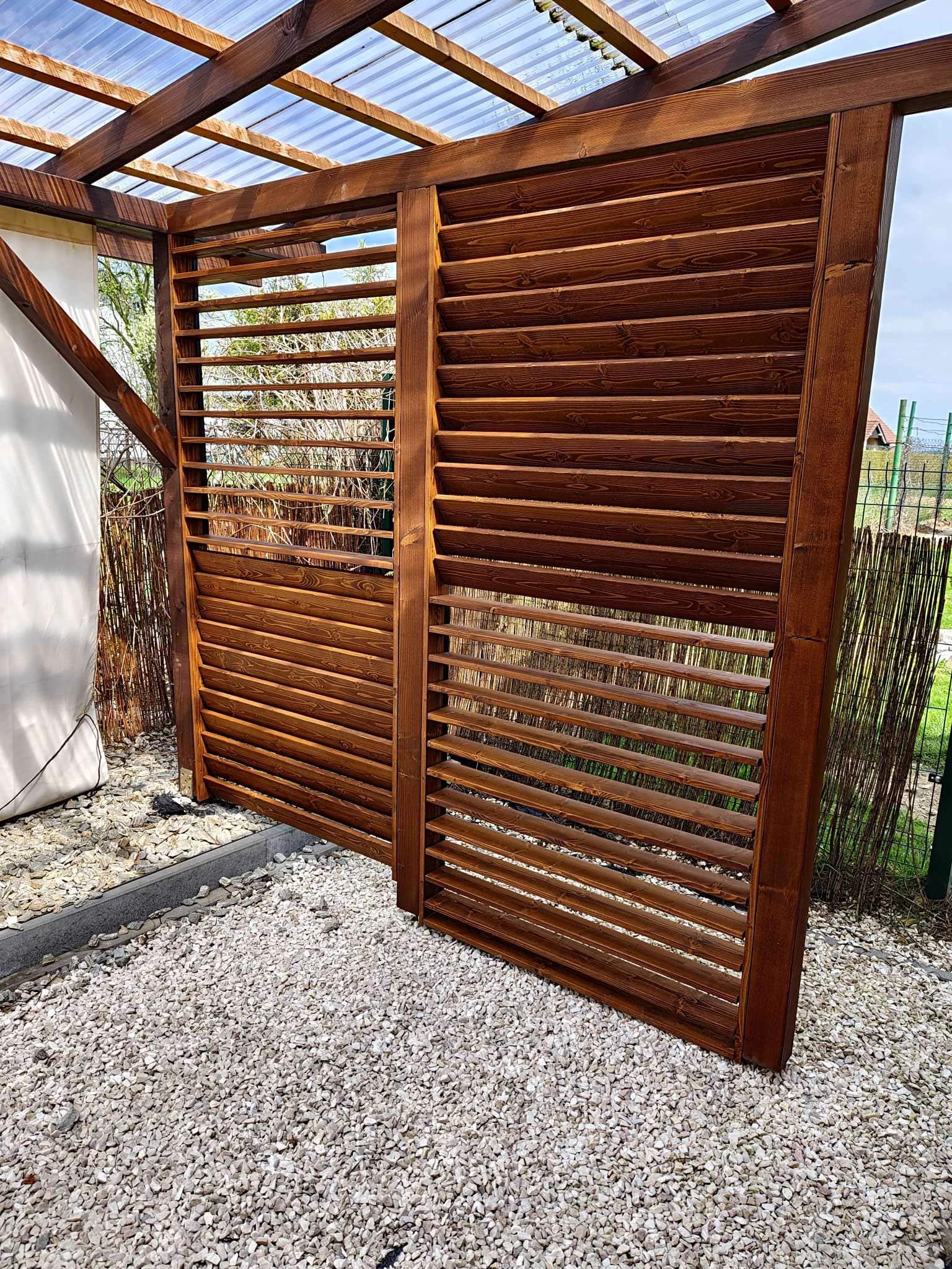 A wooden privacy screen with horizontal louvers installed outdoors on a gravel surface.