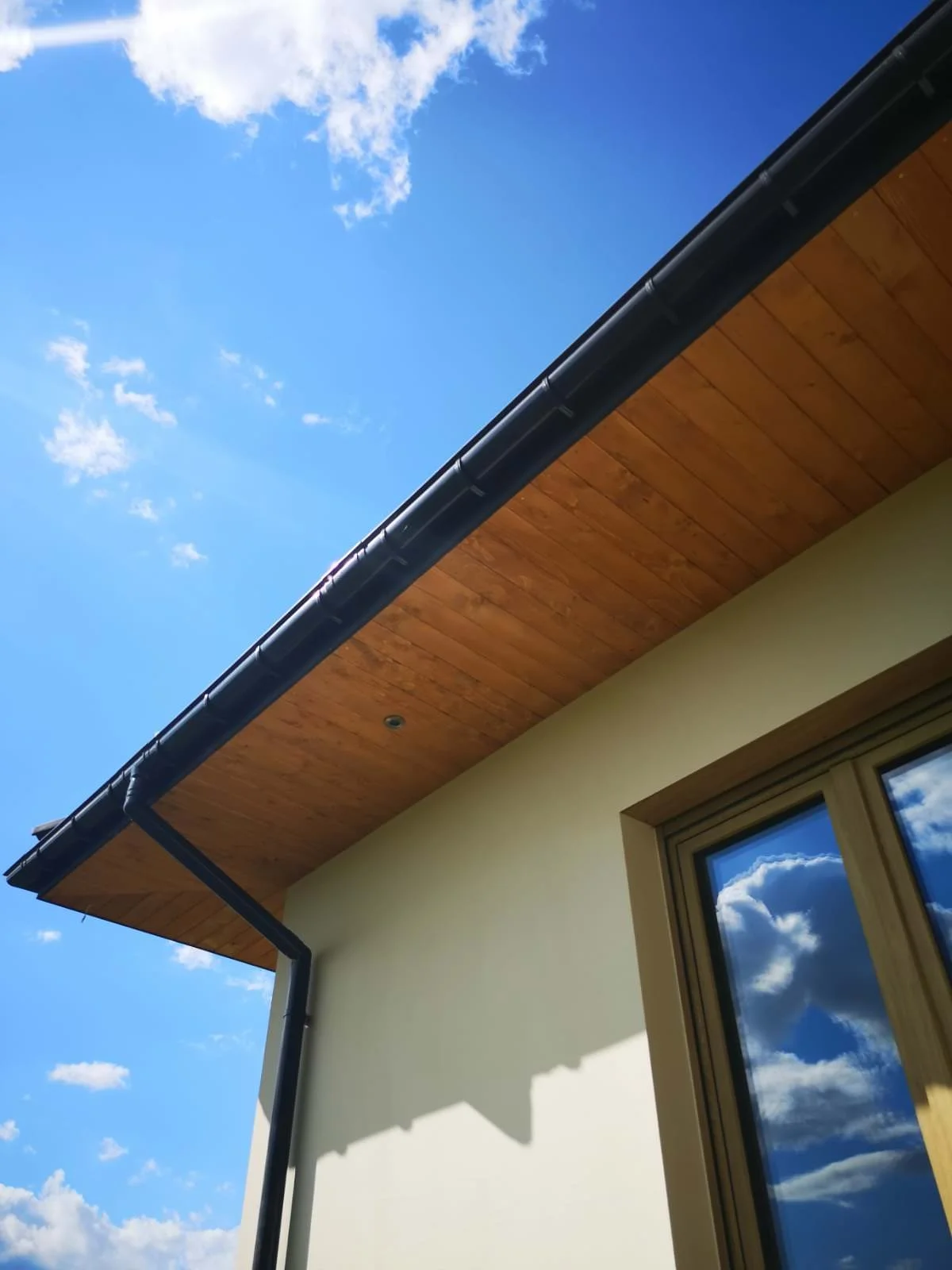 Part of a house with beige walls, a wooden roof extension, a window with a reflection of the sky and clouds, and a black gutter on a bright, partly cloudy day.