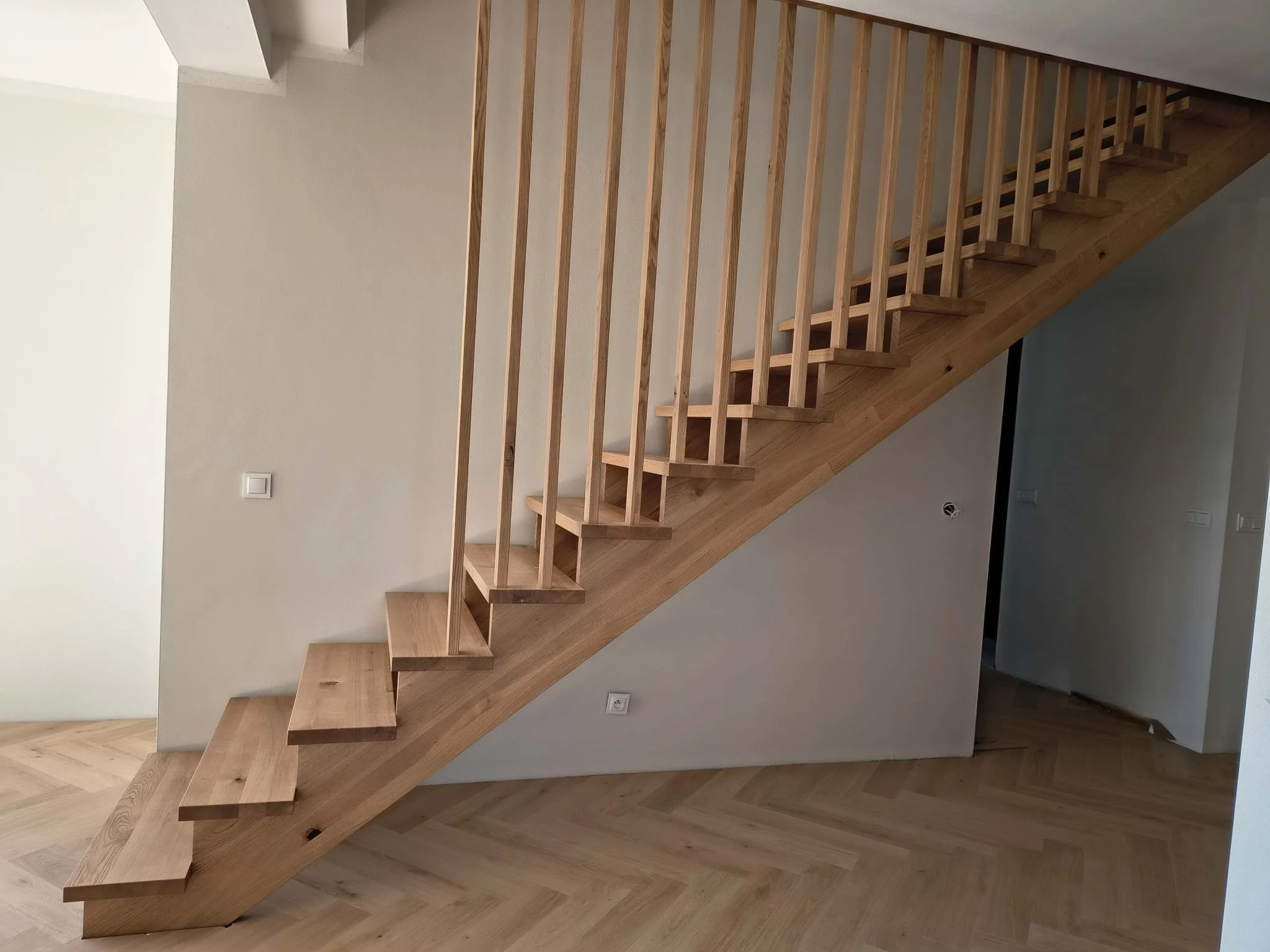 An interior view of a wooden staircase with open risers, leading upward, against a white wall with a light switch and electrical outlet nearby.