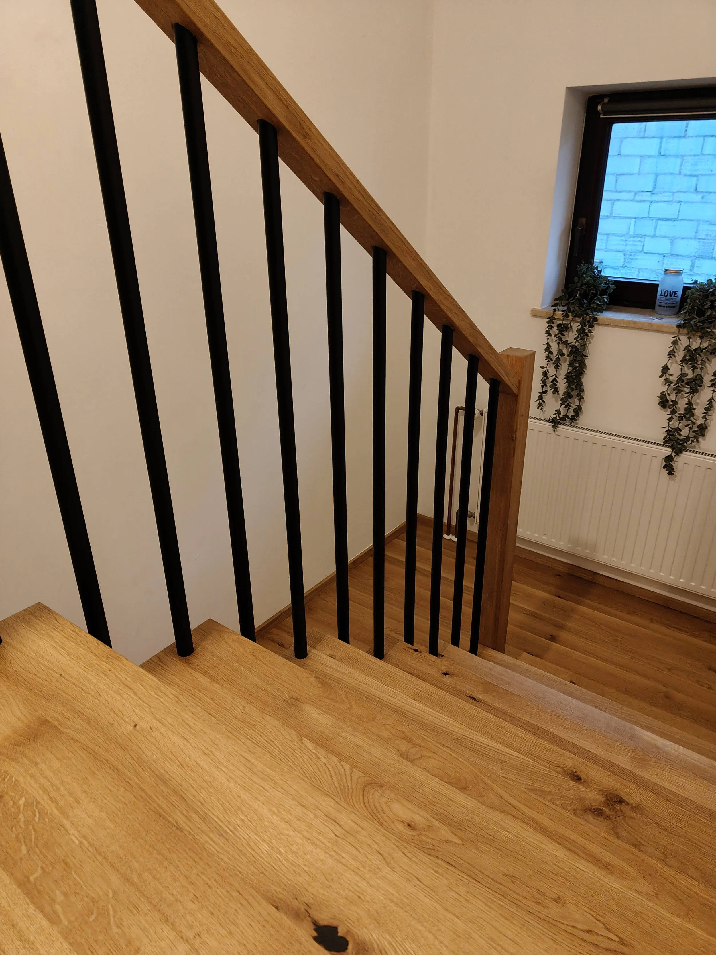 Interior view of a staircase with wooden steps, black metal balusters, a wooden handrail, and a window with a brick wall outside. There are decorative plants and a jar with the word 'LOVE' on the window sill, and a radiator below the window.