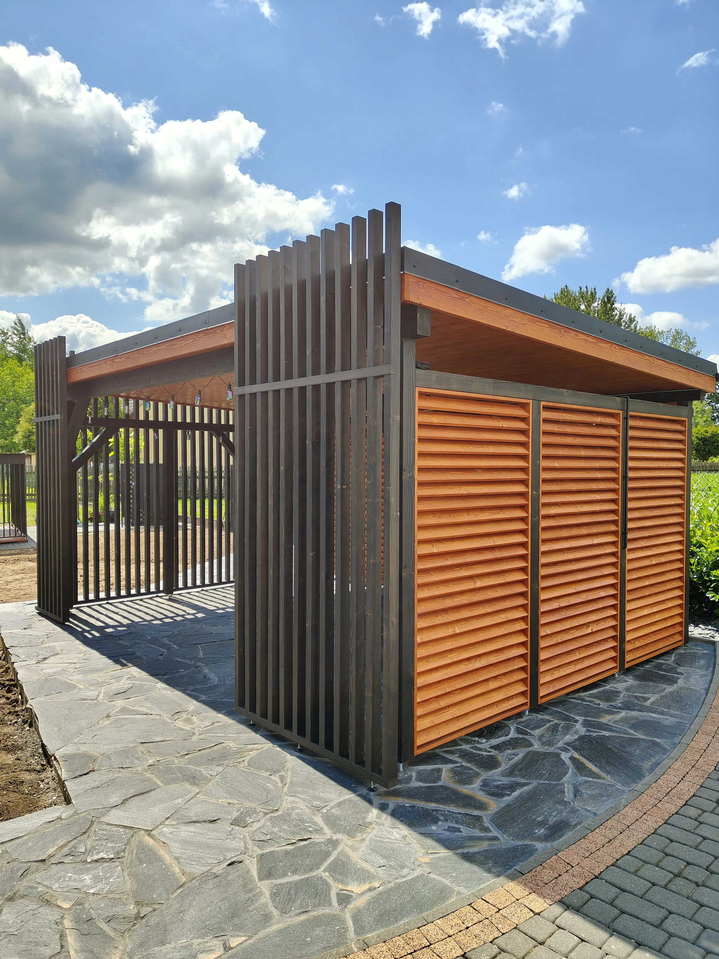 A modern outdoor structure with black vertical wooden slats and a roof with orange-brown horizontal slats, situated on a stone patio with a colorful brick border. Bright sky with clouds is visible in the background.