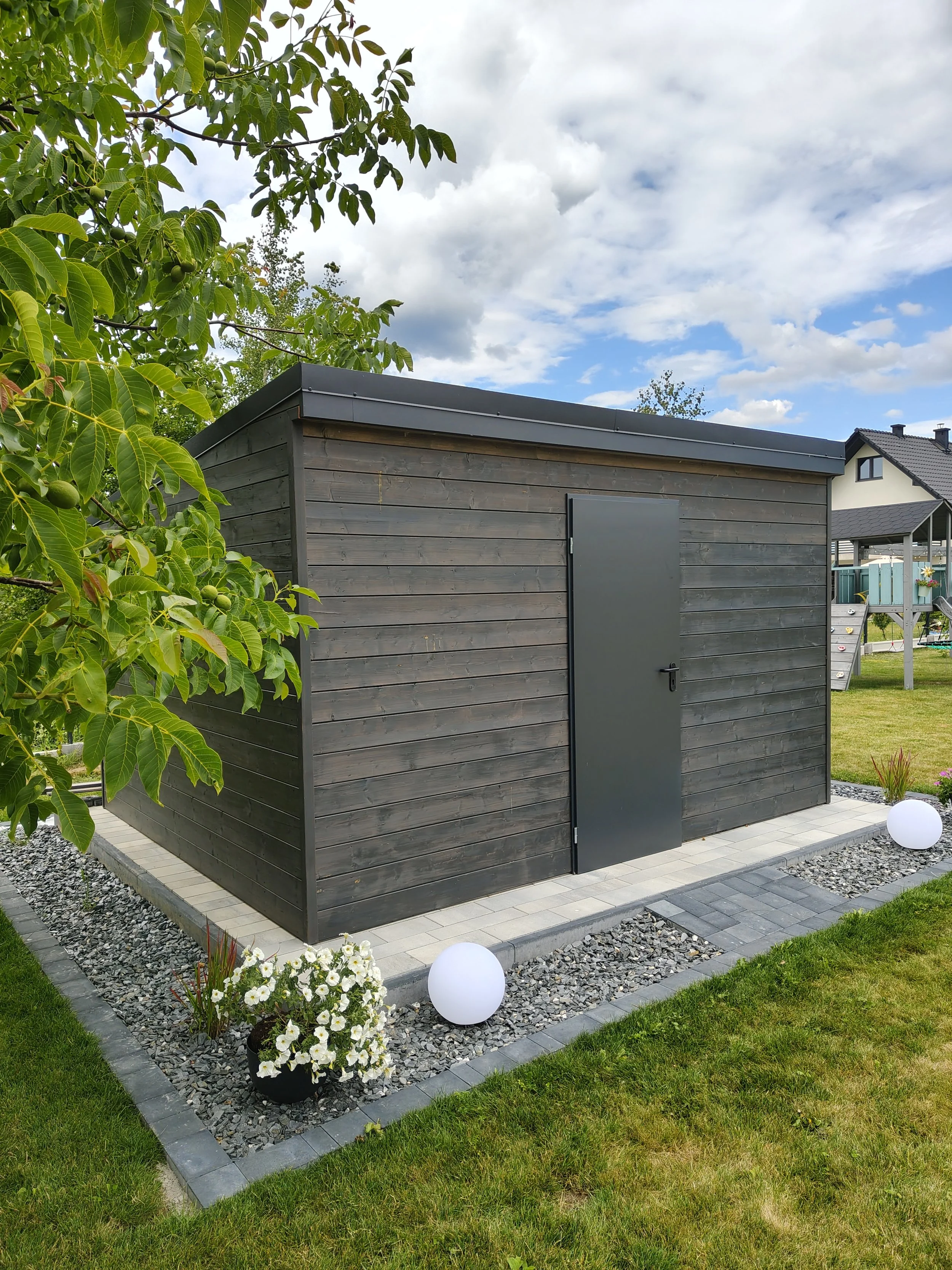 A small modern wooden shed painted dark gray, situated in a neatly landscaped yard with grass, a flower bed, and decorative white globes outside.