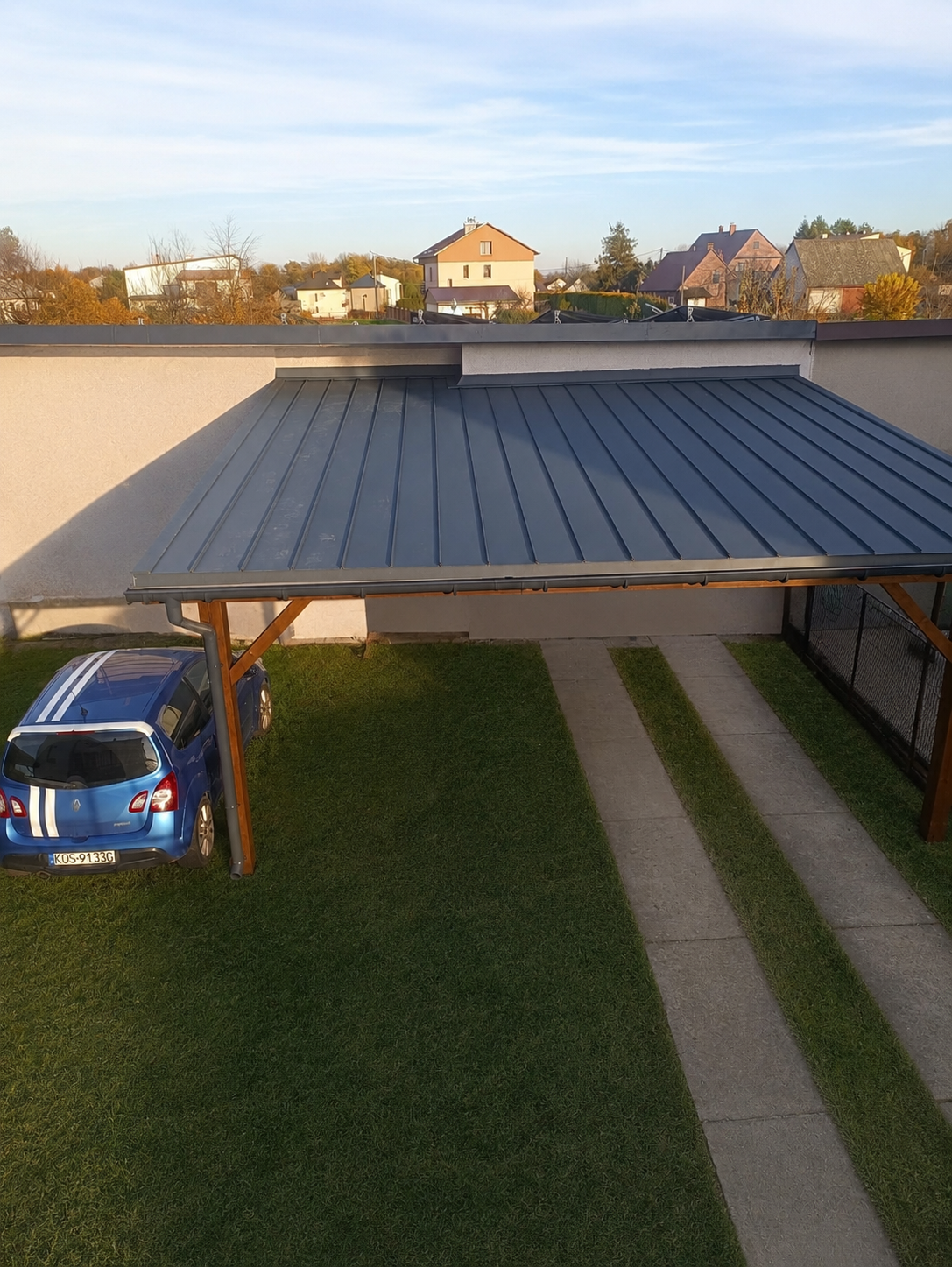 A blue car parked under a metal-roofed carport with concrete driveway strips on a grassy lawn in a residential area with houses and trees in the background.