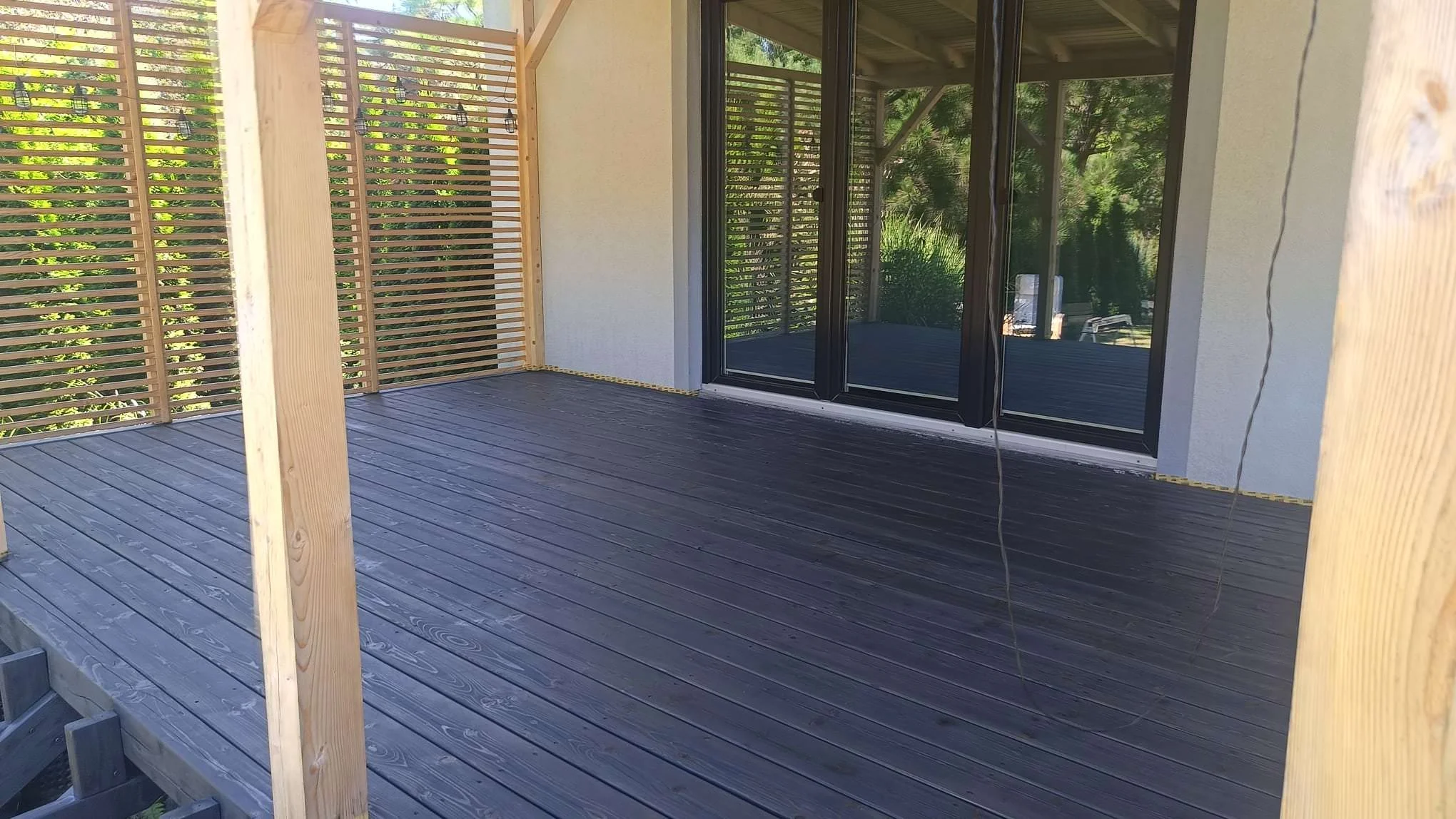 A newly built outdoor wooden deck with railings, situated in a backyard with greenery, adjacent to a house with black sliding glass doors.
