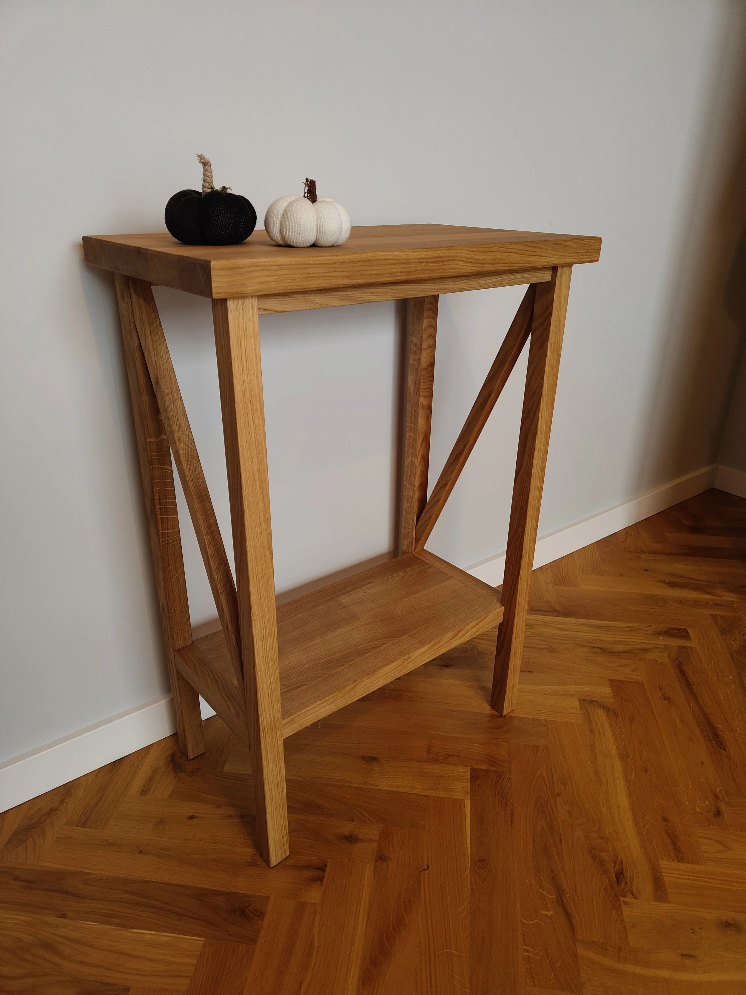 Wooden shelf with two decorative pumpkins, one black and one white, on top of it, against a light-colored wall and hardwood floor.