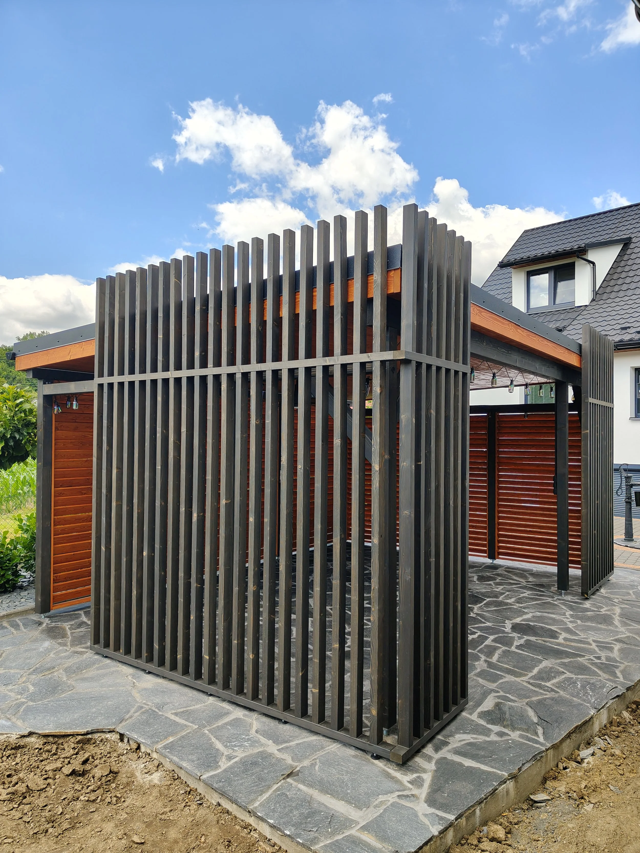 Modern outdoor structure with vertical wooden slats and a stone patio, attached to a house with a dark pitched roof and dormer window, under a partly cloudy blue sky.
