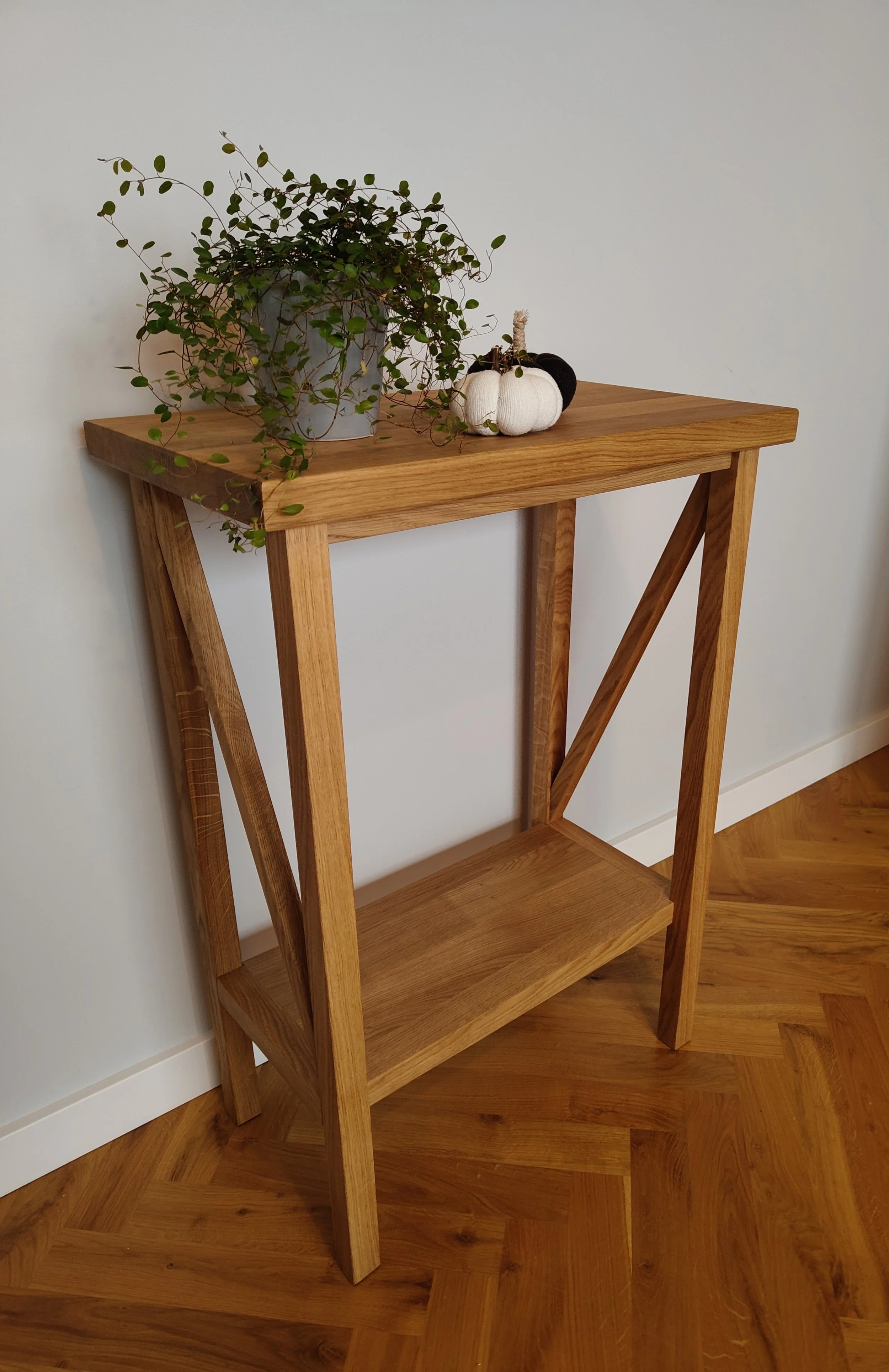 A wooden table with a decorative arrangement of white and black fabric pumpkins and a potted trailing green plant on top, positioned against a plain white wall.