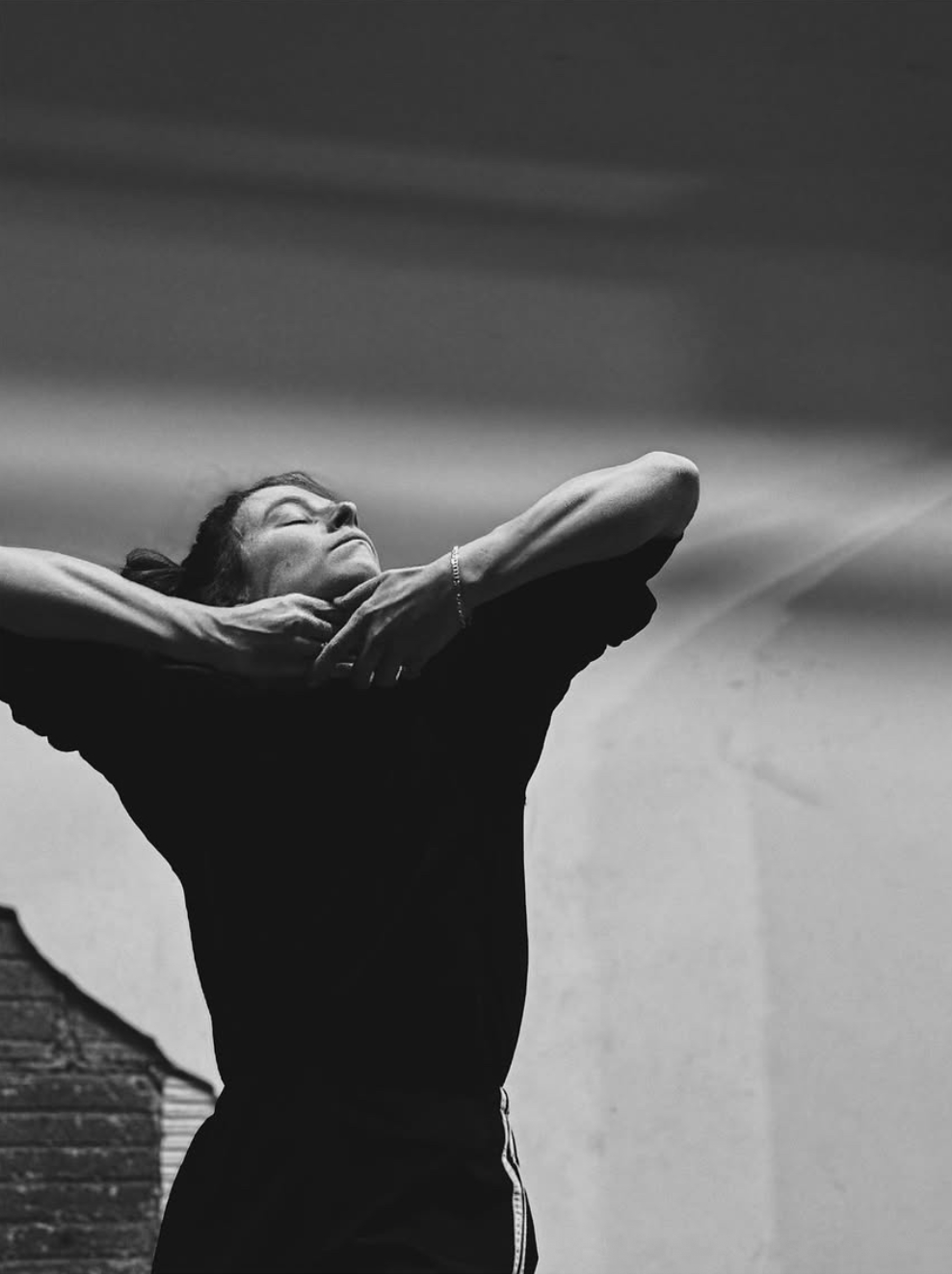 A woman with dark hair, eyes closed, appears to be stretching or relaxing with arms raised behind her head, in a black and white photo.