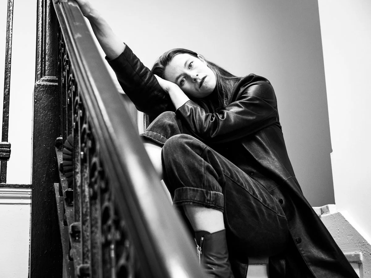 Black and white photo of a woman sitting on stairs, resting her head on her arm, wearing a leather jacket and jeans, looking at the camera.