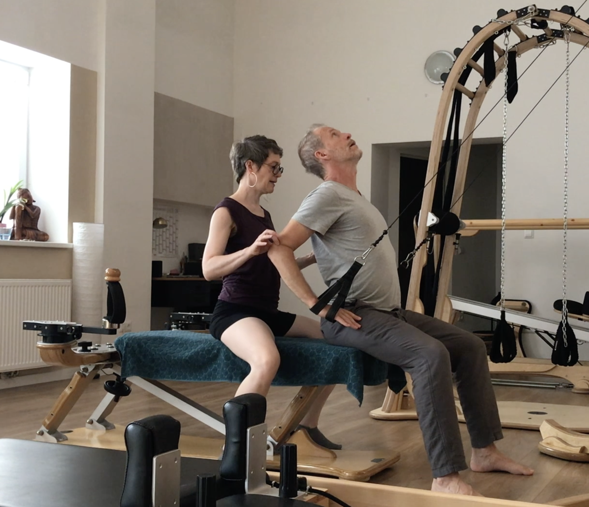 A woman and a man are engaging in a session. The woman is standing behind the man, holding his arms and guiding him as he leans back into the inversion. They are in a well-lit room with modern decor, with a statue and some decor on a shelf visible in the background.