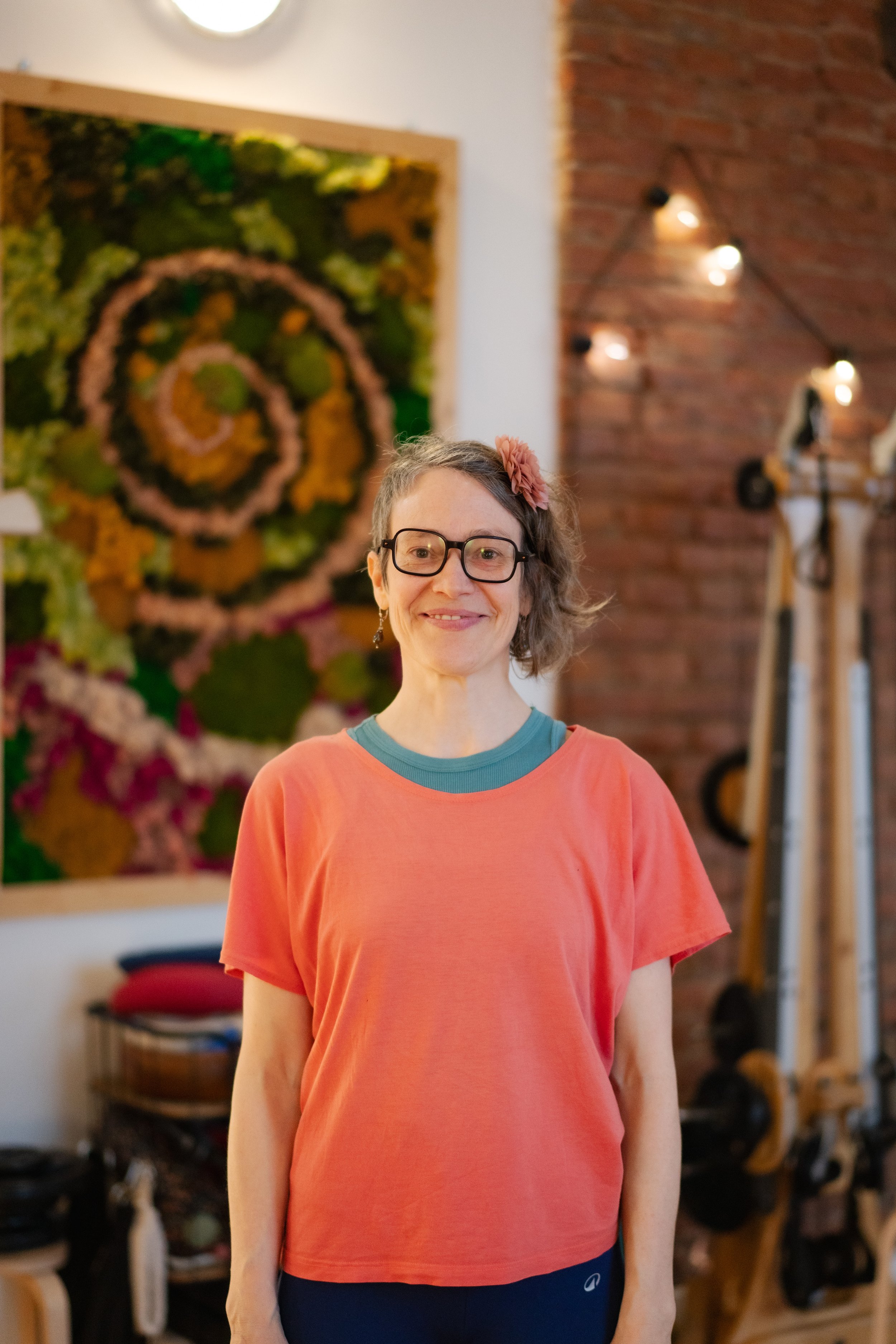 A woman with glasses and a pink flower in her hair, smiling, wearing an orange t-shirt, standing inside a room with Gyrotonic® exercise equipment and artwork on the wall.