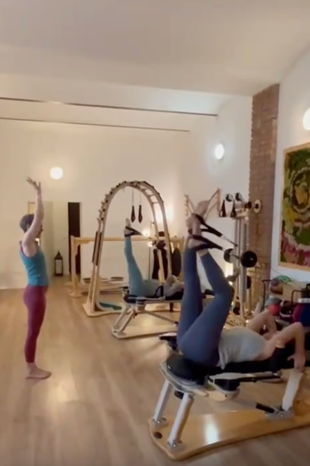 Three people engaged in a Gyrotonic® class in a room with wooden flooring, white walls, a brick accent wall, and modern lighting. The trainer is standing with arms raised, the two clients are lying on exercise equipment.