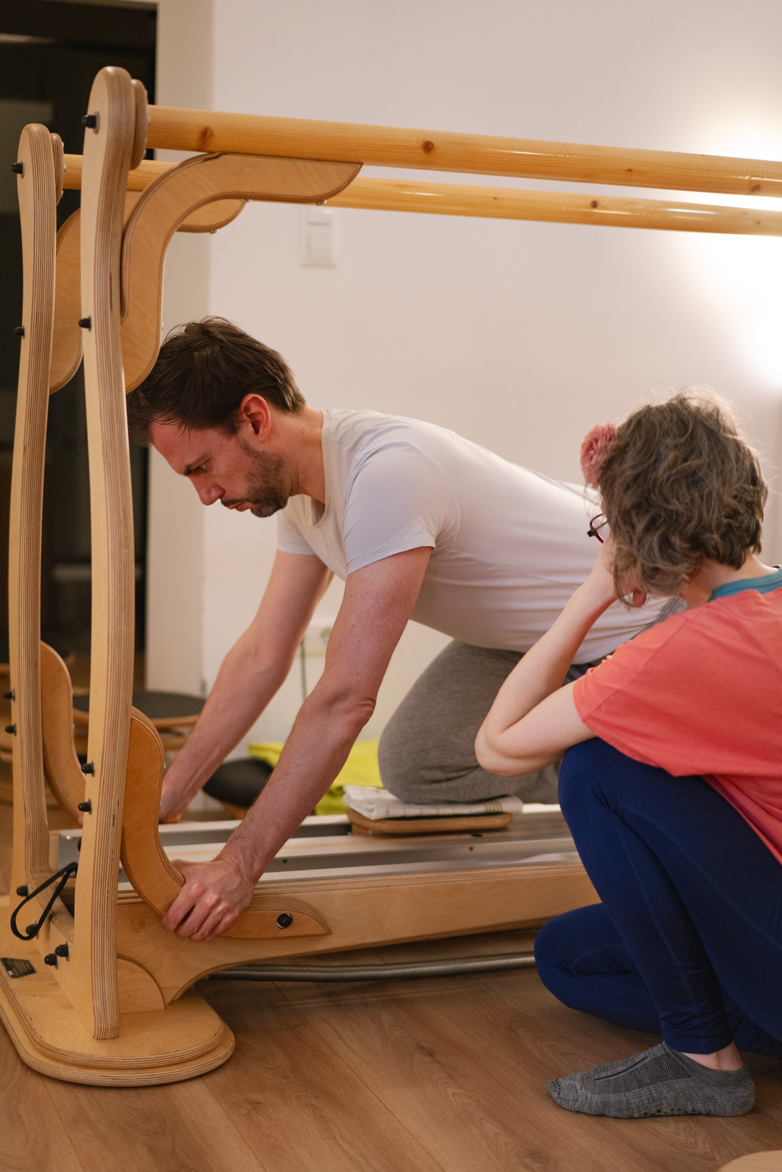A client in a Gyrotonic class using the Jumping Stretching Board equipment. The client is kneeling and leaning forward, with his hands on the machine, while the trainer is kneeling next to him to give instructions and hands on.