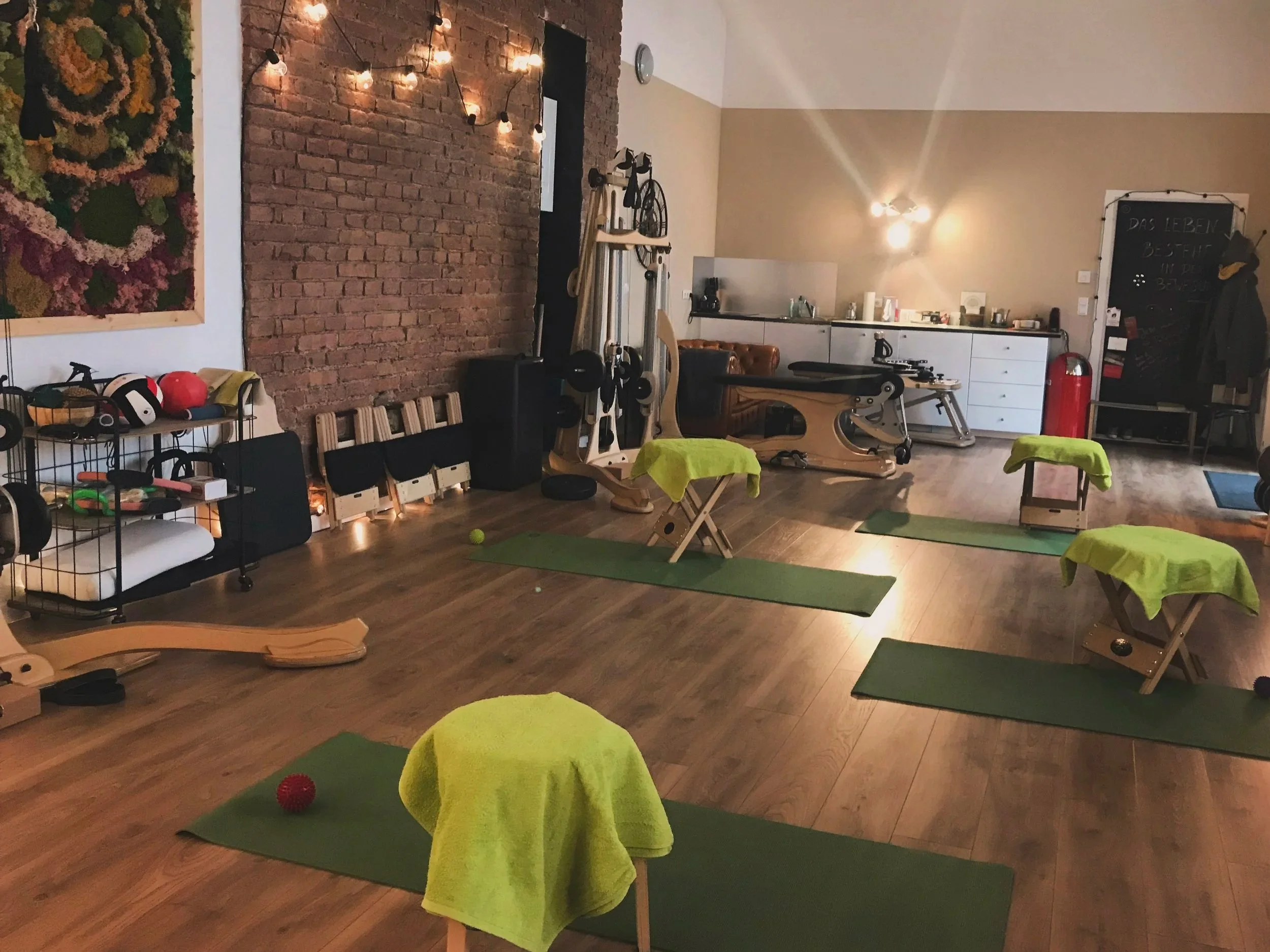 Interior of a Gyrotonic studio with green yoga mats, Gyrotonic stools, exercise equipment, and wall decorations including a large colorful floral artwork. There are string lights on a brick wall, and a chalkboard with writing in German.