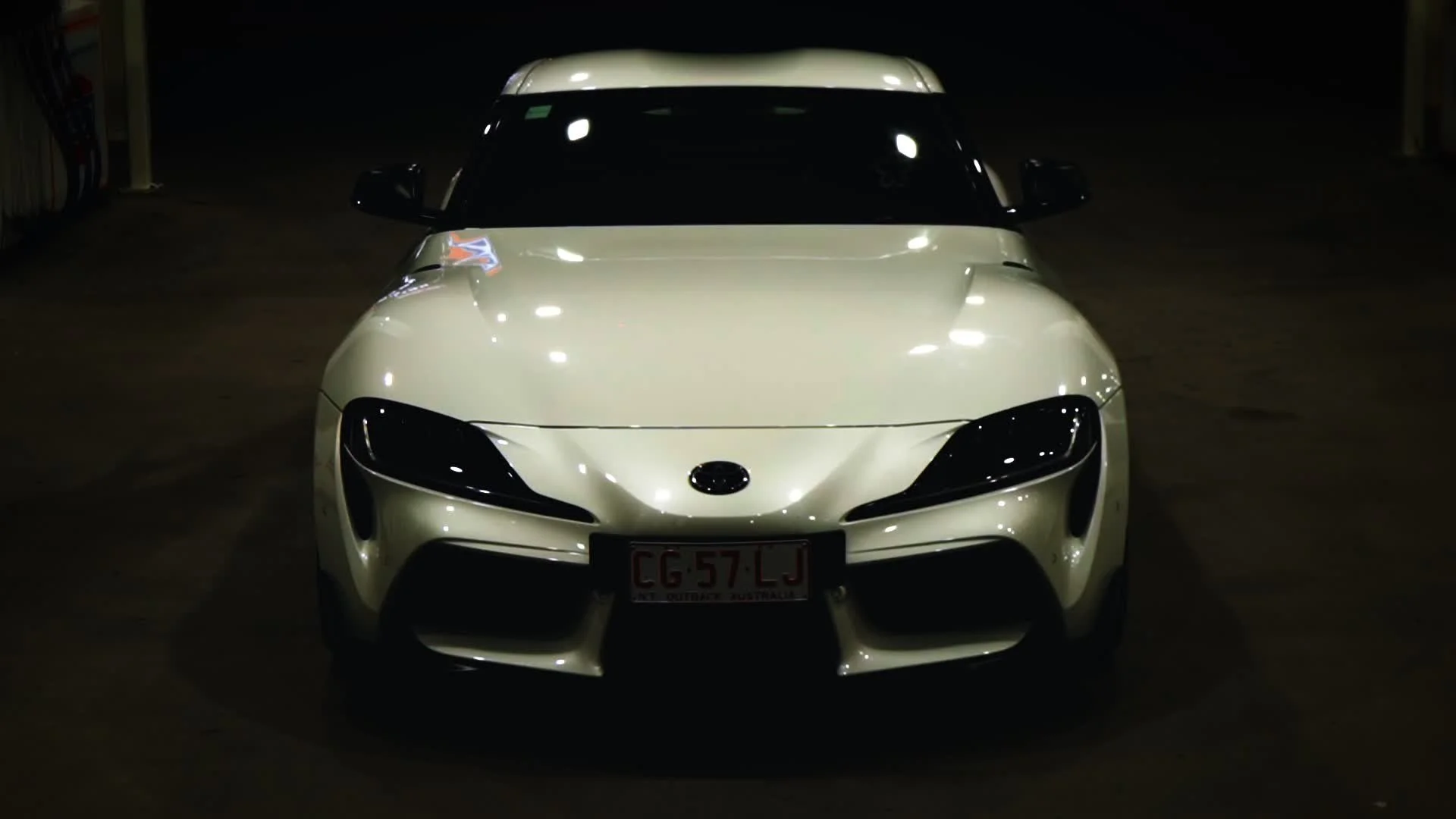 Front view of a white sports car parked in a dark garage, with black headlights and a license plate reading "GC57LJ".