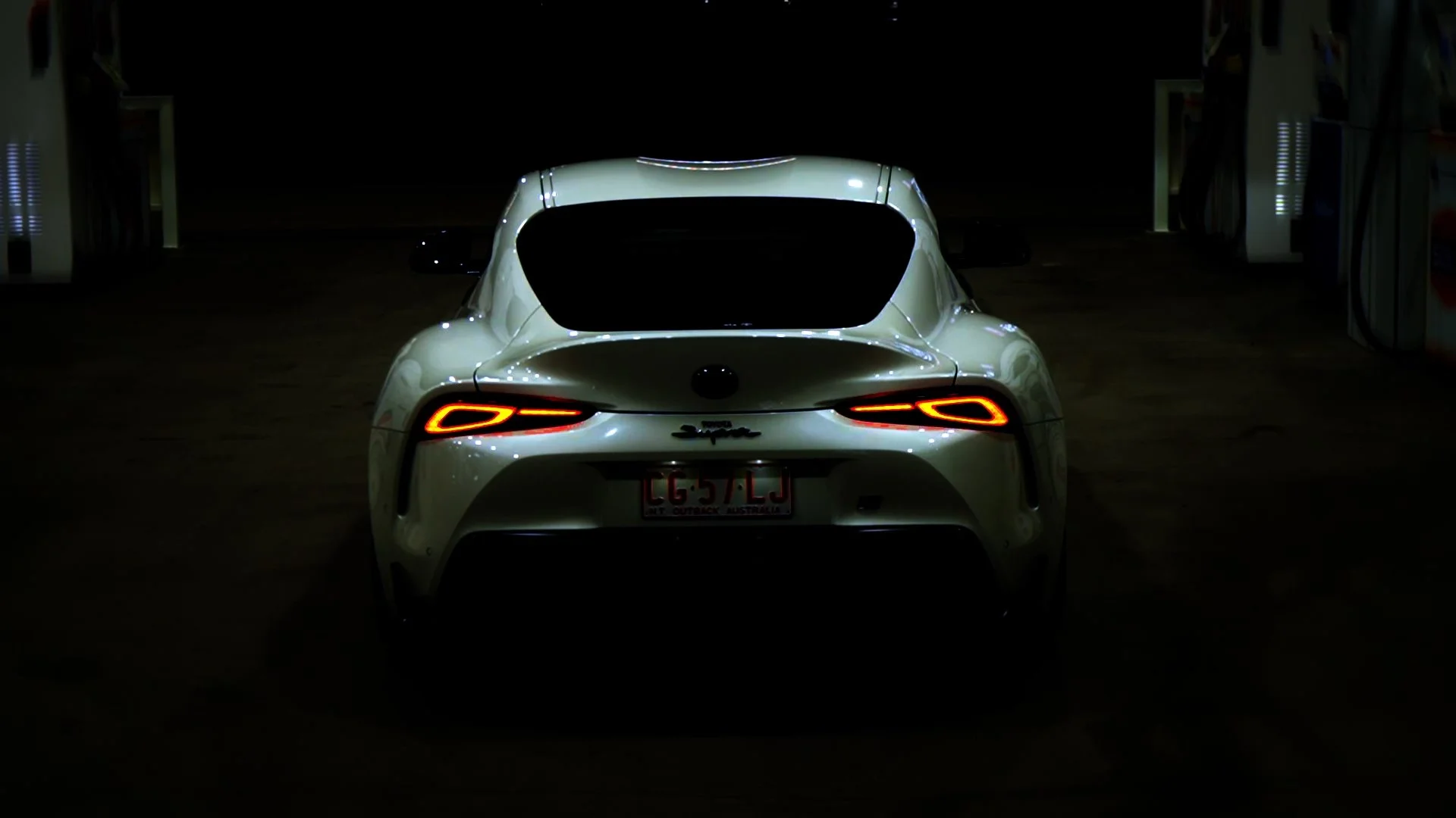 White luxury sports car parked in a dimly lit garage with illuminated tail lights and license plate.
