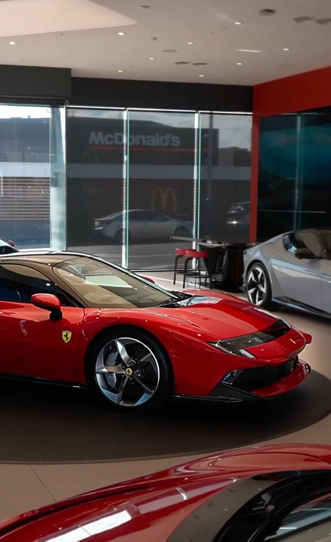 Red Ferrari sports car inside a showroom with other cars and large windows showing a McDonald's restaurant outside.