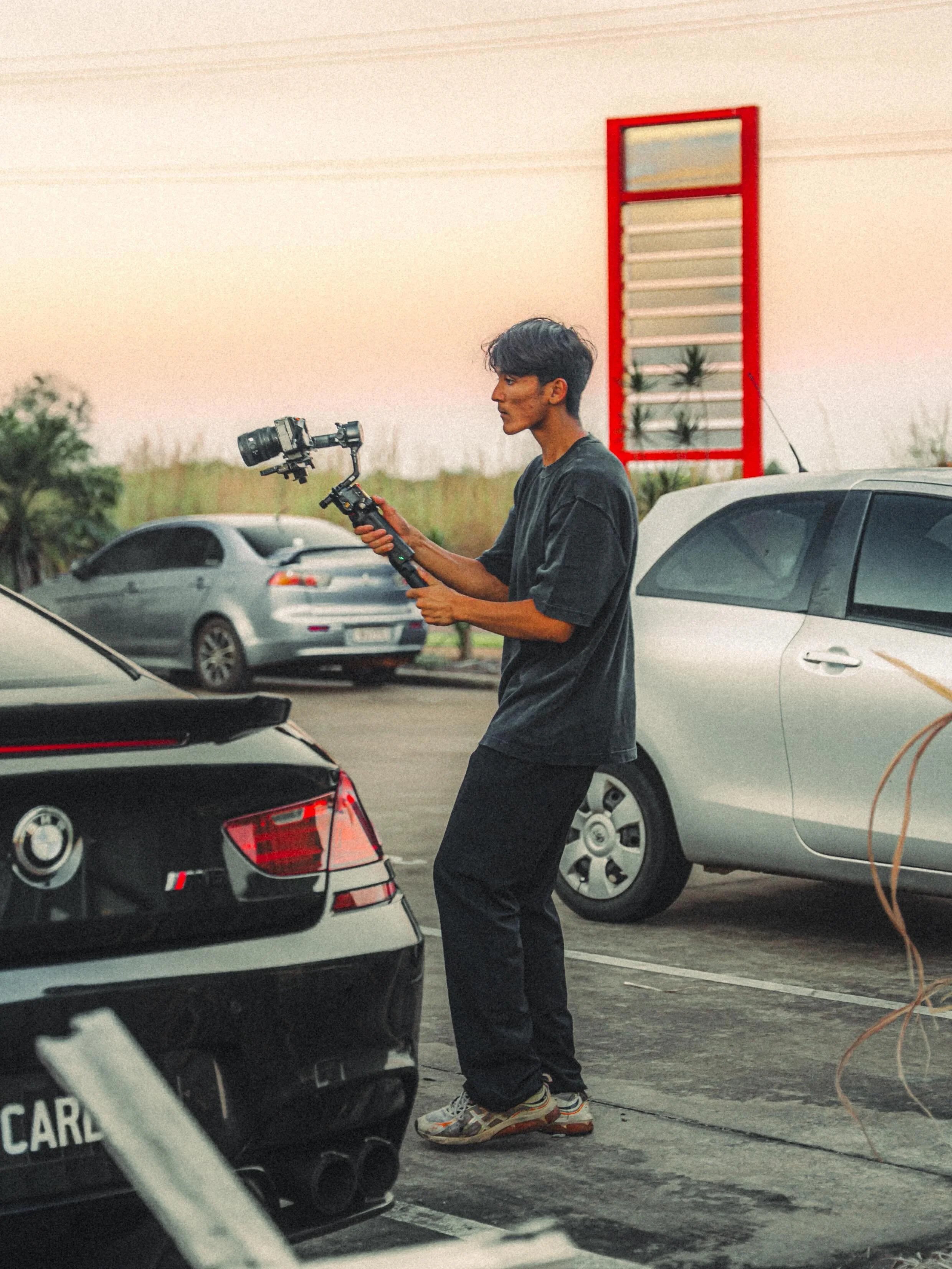 Young man standing in parking lot, holding a camera gimbal, wearing black oversized t-shirt, black pants, and sneakers, with cars parked around him.