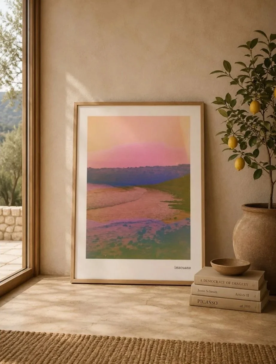 Framed colorful landscape art print leaning against a wall next to a potted lemon tree, some books, and a small bowl, near a window with sunlight streaming in.