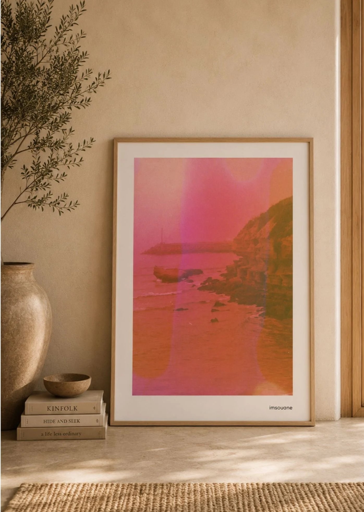 A framed pink and orange landscape painting leaning against a beige wall, with a tall vase with greenery on the left, a small bowl, and a stack of books on the bottom left corner, atop a light-colored surface. The books have titles such as "Kinfolk," "Hide and Seek," and "A Life Less Ordinary."