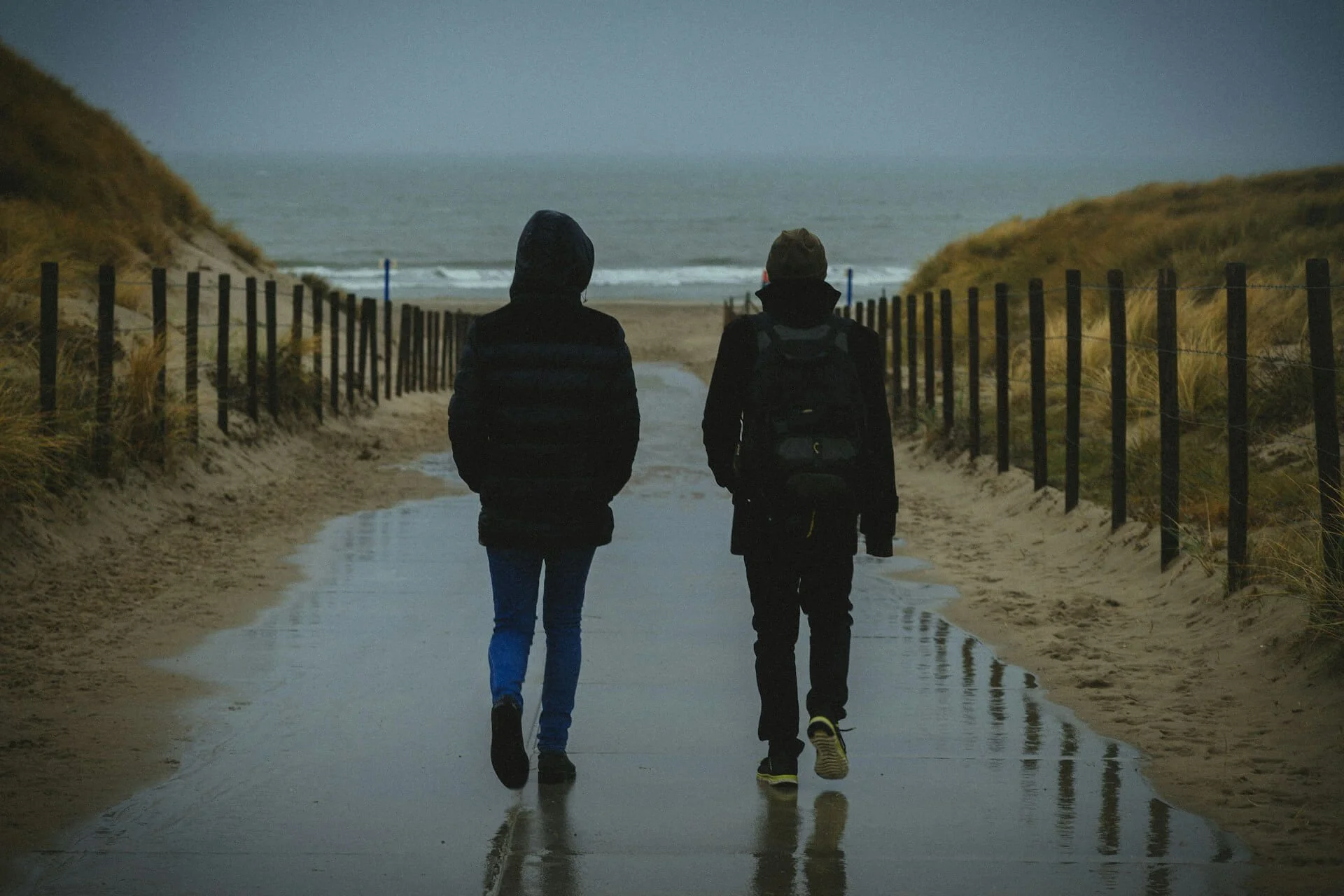 two people with thick warm jackets walking along a wet path leading to waves on a beach