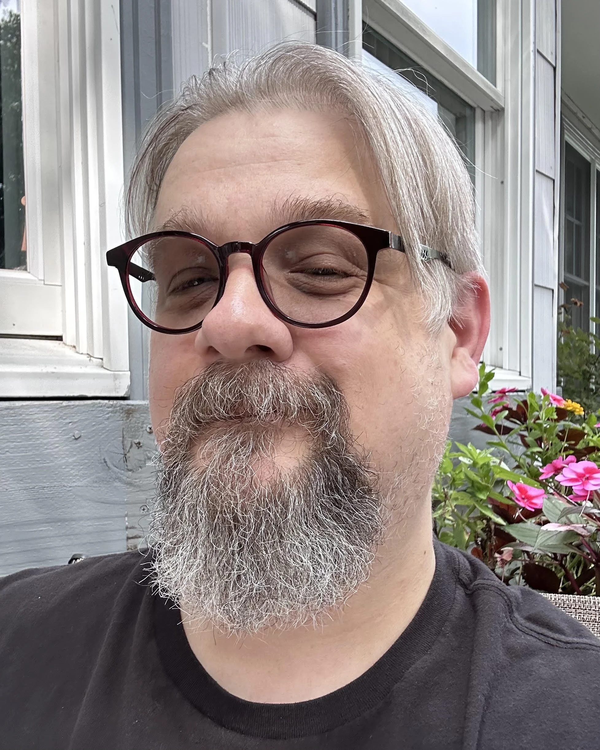 A man with light-colored hair, glasses, and a beard smiling outdoors near a house with windows and flowers.