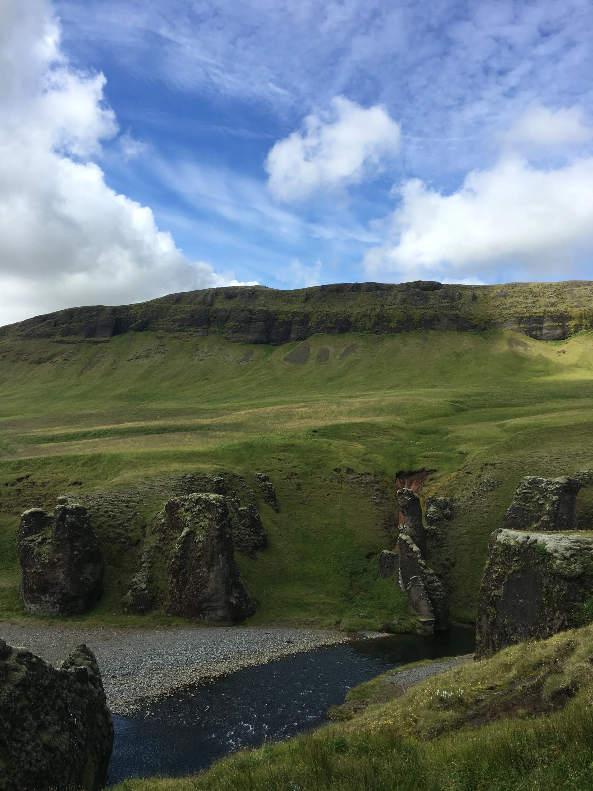 A river flowing through a lush green valley with moss-covered rocks in the foreground and a grassy hillside with a cliff in the background under a partly cloudy sky.