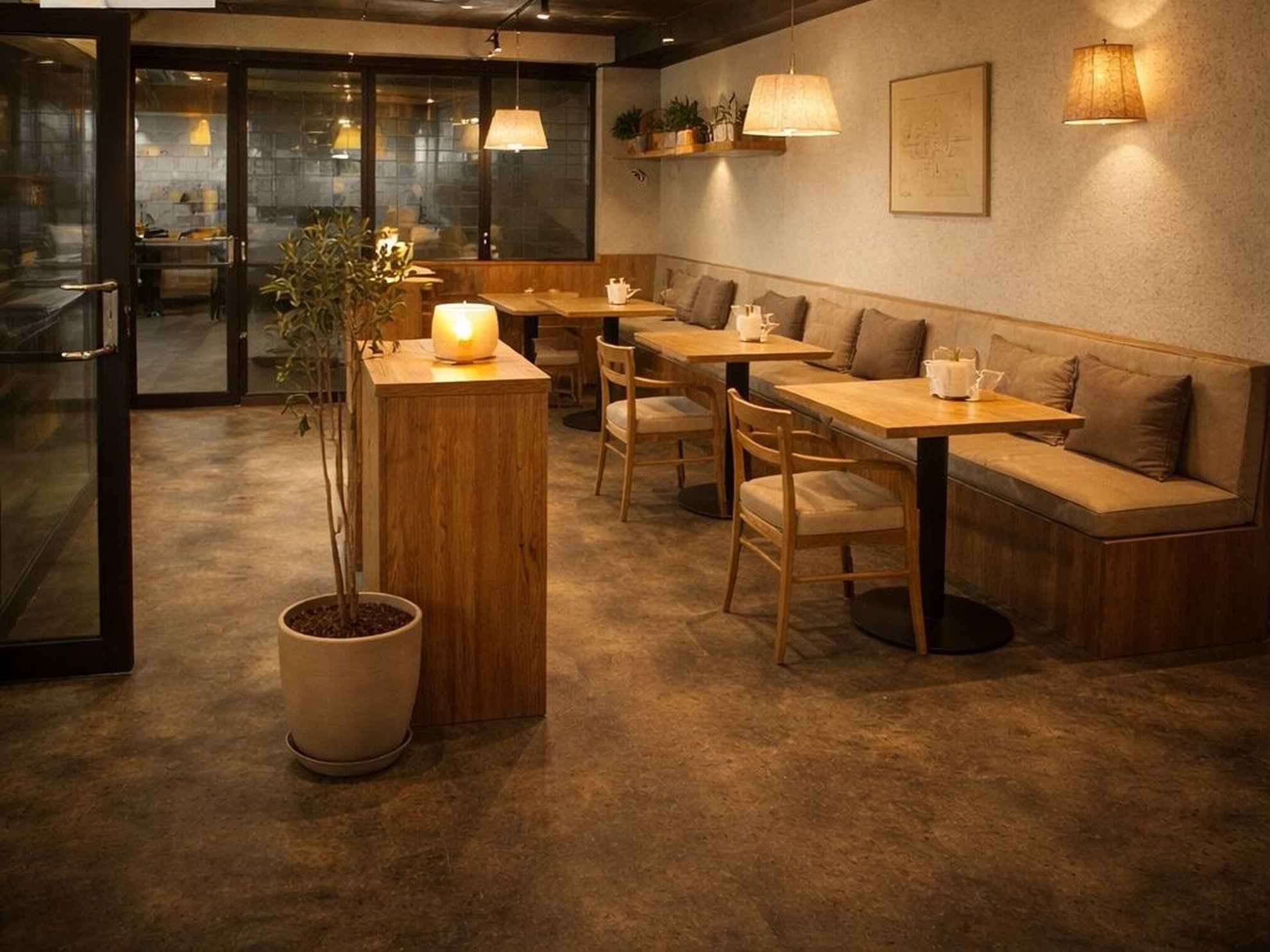 Warm and cozy restaurant interior with wooden tables and chairs, cushioned bench seating, hanging pendant lights, potted plant, decorative shelves with plants, framed artwork on the wall, and a lit candle on a wooden counter.