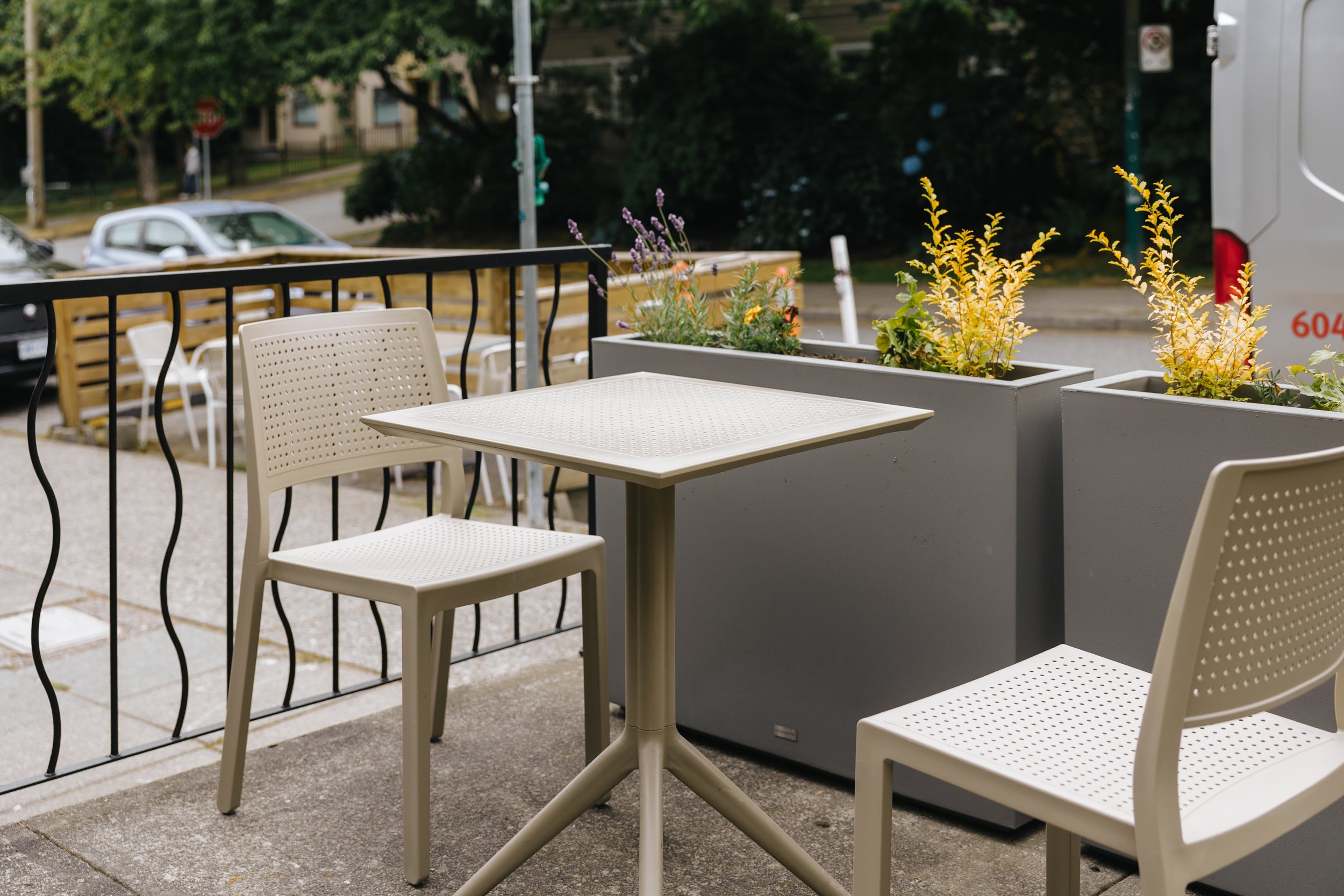 Outdoor patio with white perforated chairs, a white table, landscaped planters with flowers, and a parking lot with cars and trees in the background.