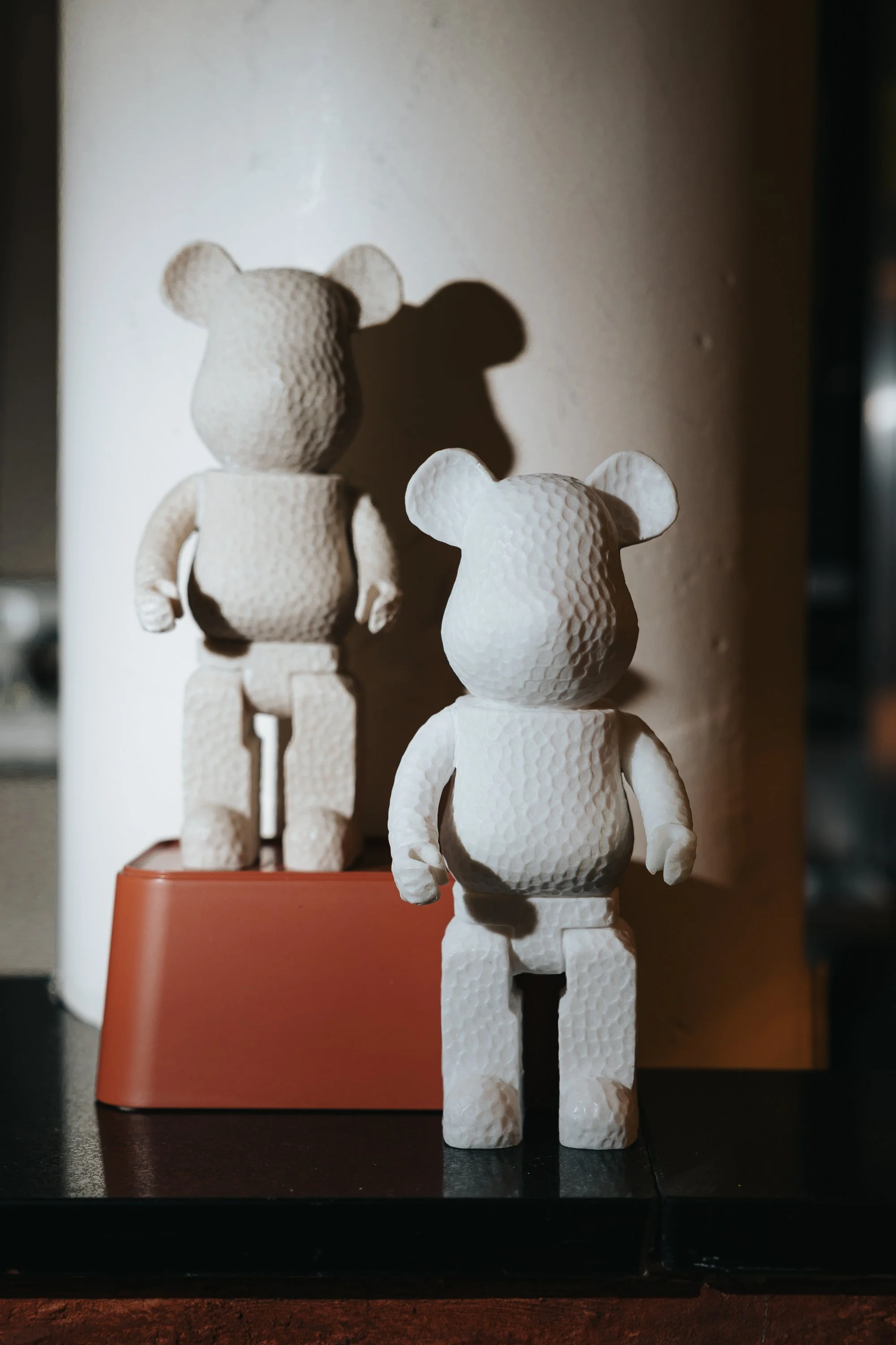 Two white teddy bear figurines with textured surfaces, one positioned behind the other, standing on a dark surface. The one in the foreground has a head with rounded ears, a small body, and arms hanging down, while the one in the background is larger with a similar shape and stands on a reddish base. A large white cylindrical object with a textured surface is in the background.