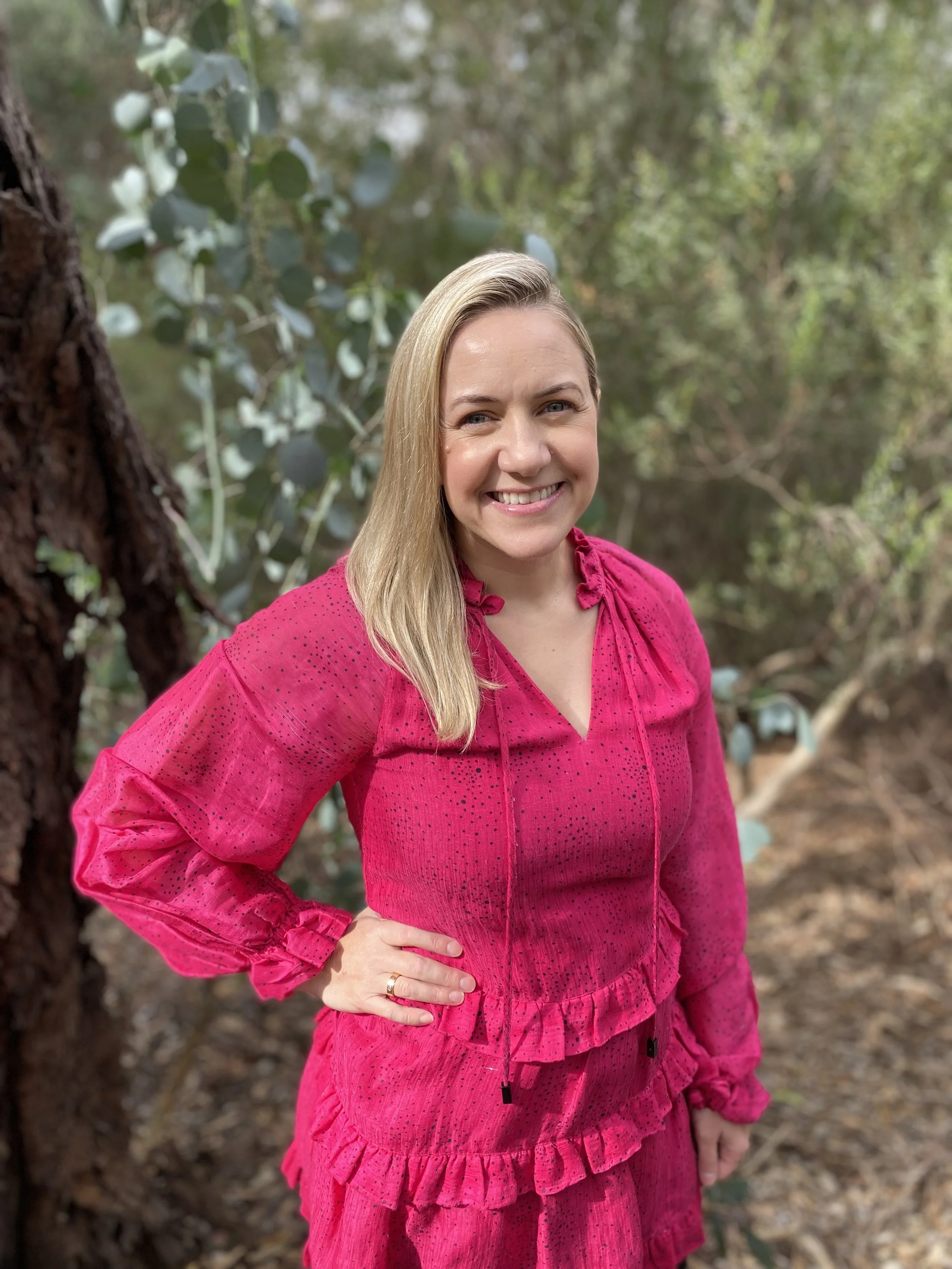 A smiling woman with long blonde hair, wearing a pink dress, standing outdoors in a wooded area.
