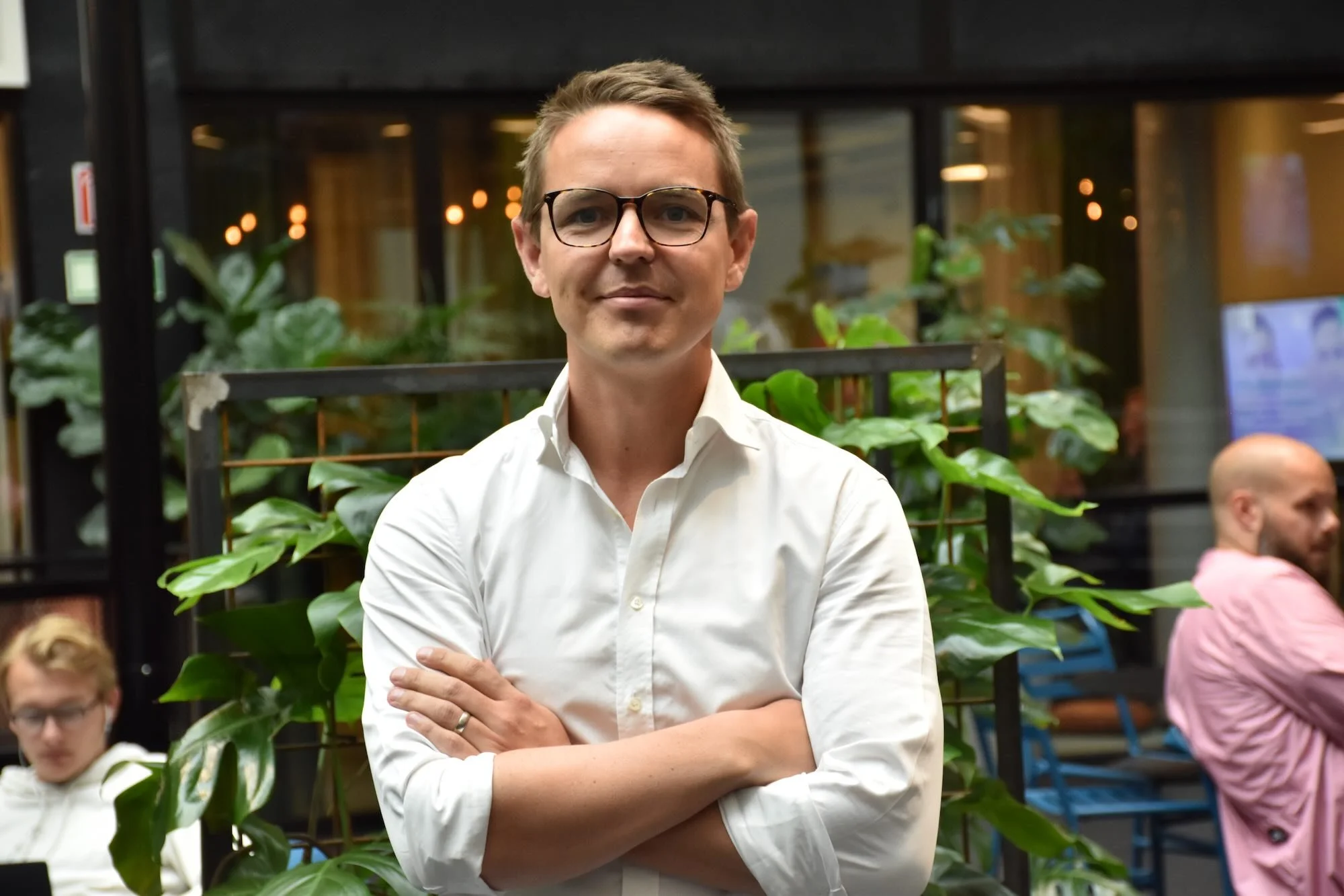 A young man with glasses and short brown hair wearing a white collared shirt, standing with his arms crossed in an indoor setting with greenery and glass walls, looking at the camera.