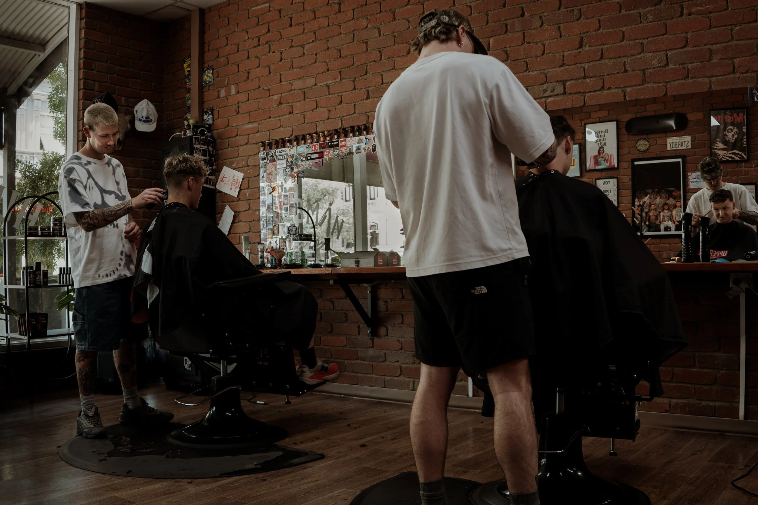 Barbershop scene with three men in a hair salon, one haircutting a customer's hair, another person standing nearby, and a third man in the background using a phone, all inside a space with brick walls and framed photos.