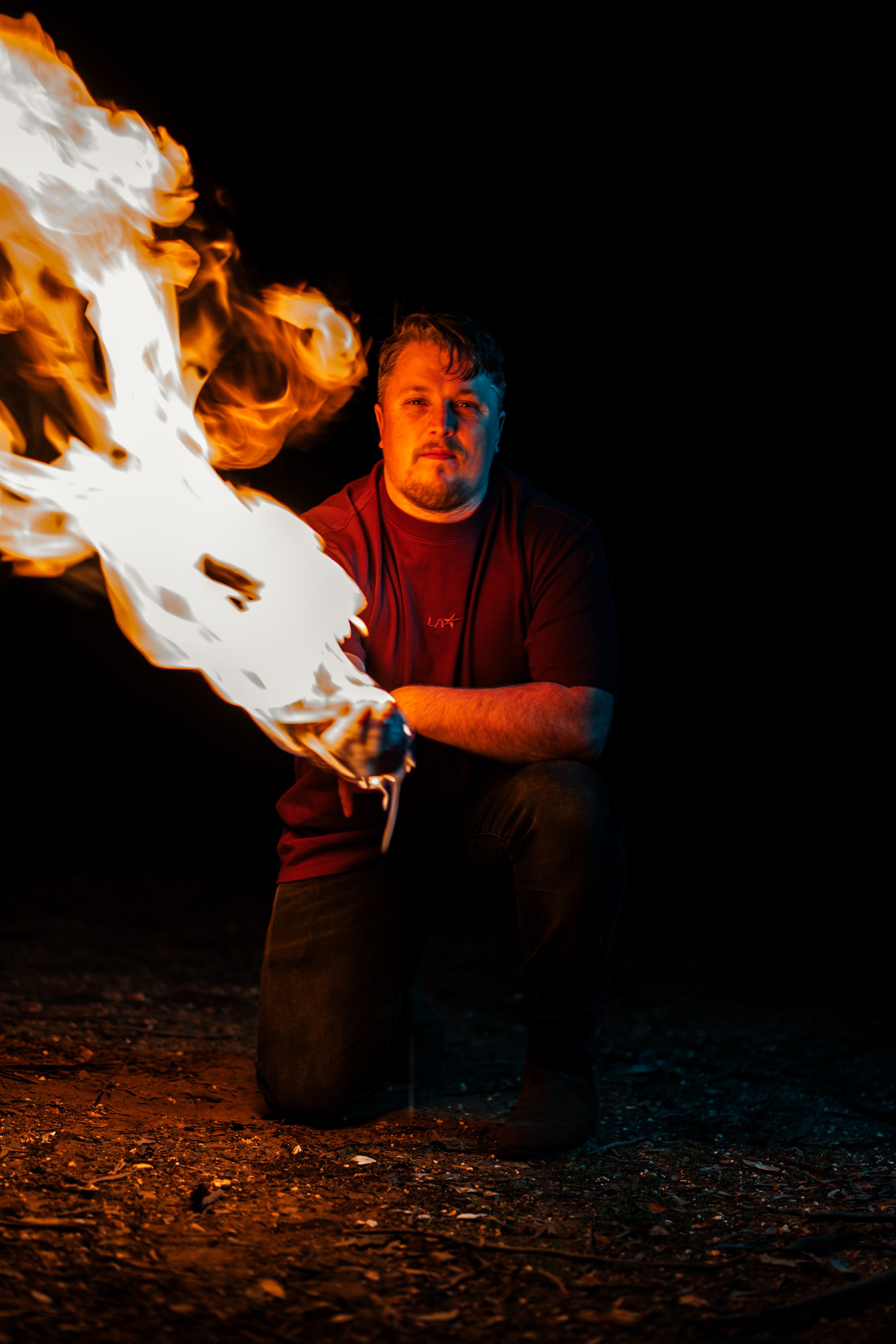 A man kneeling on the ground at night with flames in front of him, illuminated by the fire, against a dark background.