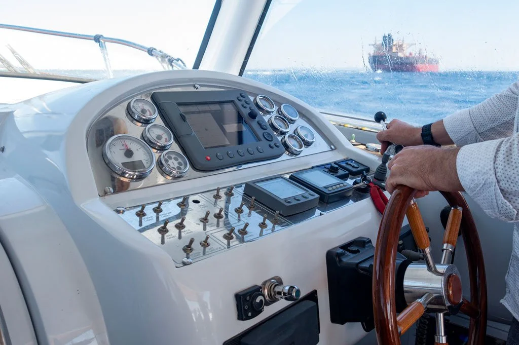 View from the cockpit of a boat showing navigational equipment, steering wheel, and a person operating the controls, with a ship in the distance at sea.