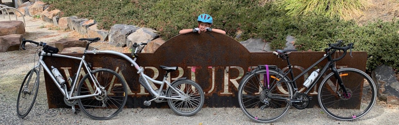 A young girl wearing a blue helmet peeking over a rusty metal sign with the word 'WARBURTON' on it, with two bicycles parked in front of the sign on a gravel path. This is the Warburton to Lilydale rail trail