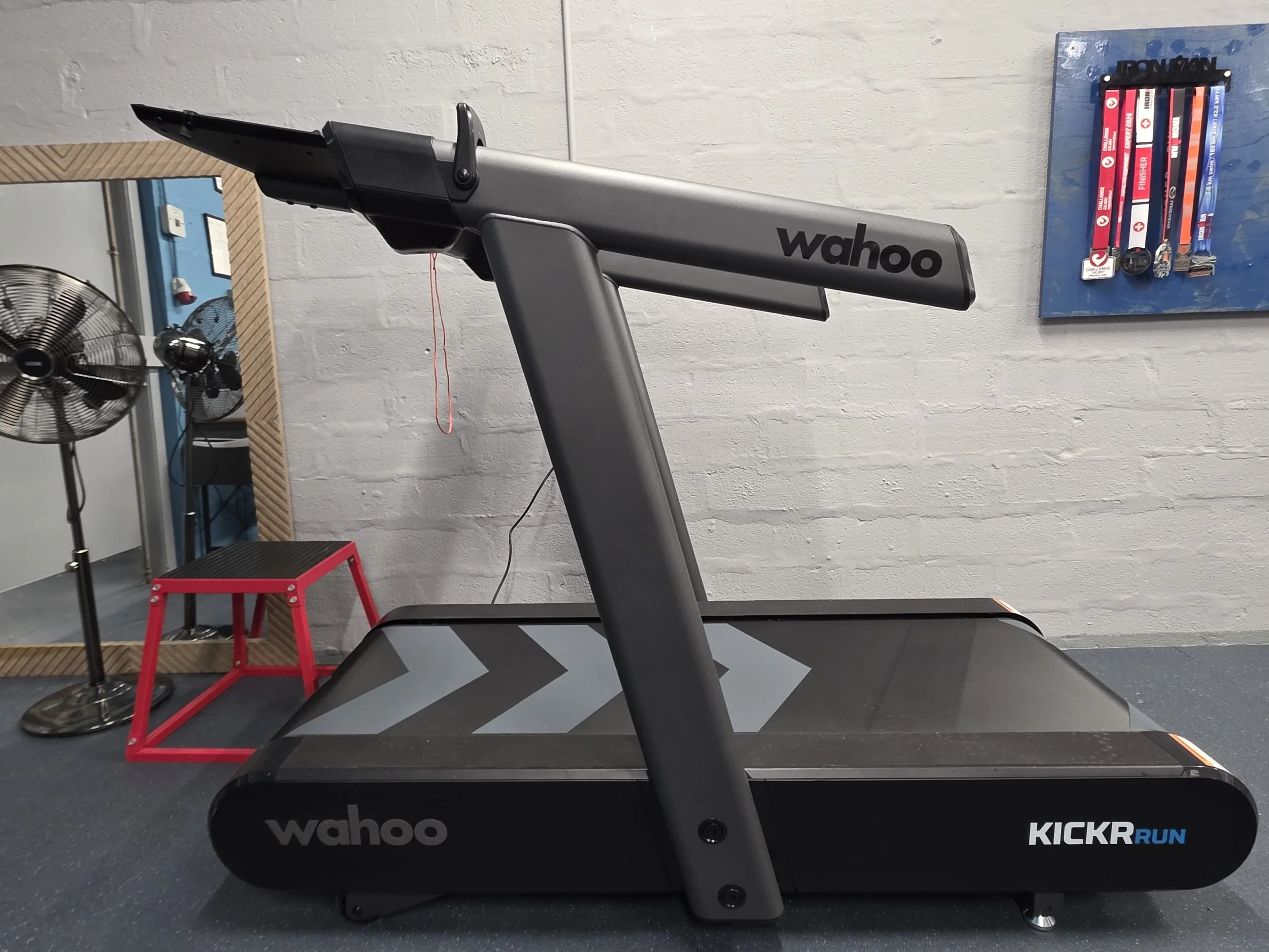 Wahoo KICKR RUN treadmill in a home gym, with a fan, step platform, mirror, and medal display.