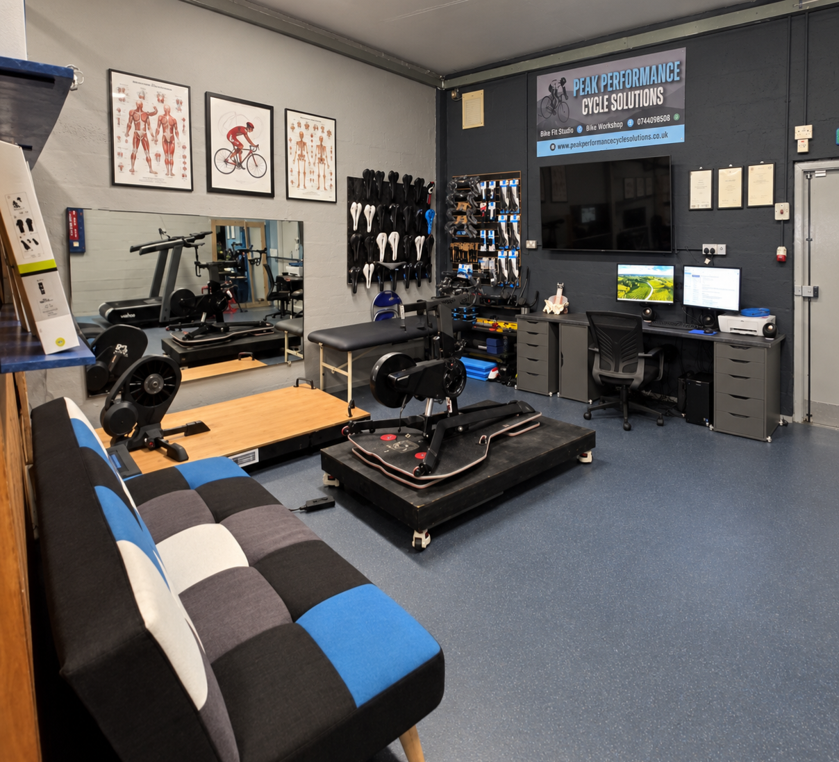 Peak Performance Cycle Solutions bicycle workshop with cycling fitness equipment, bike saddles, tools, anatomy posters, and computer workstation