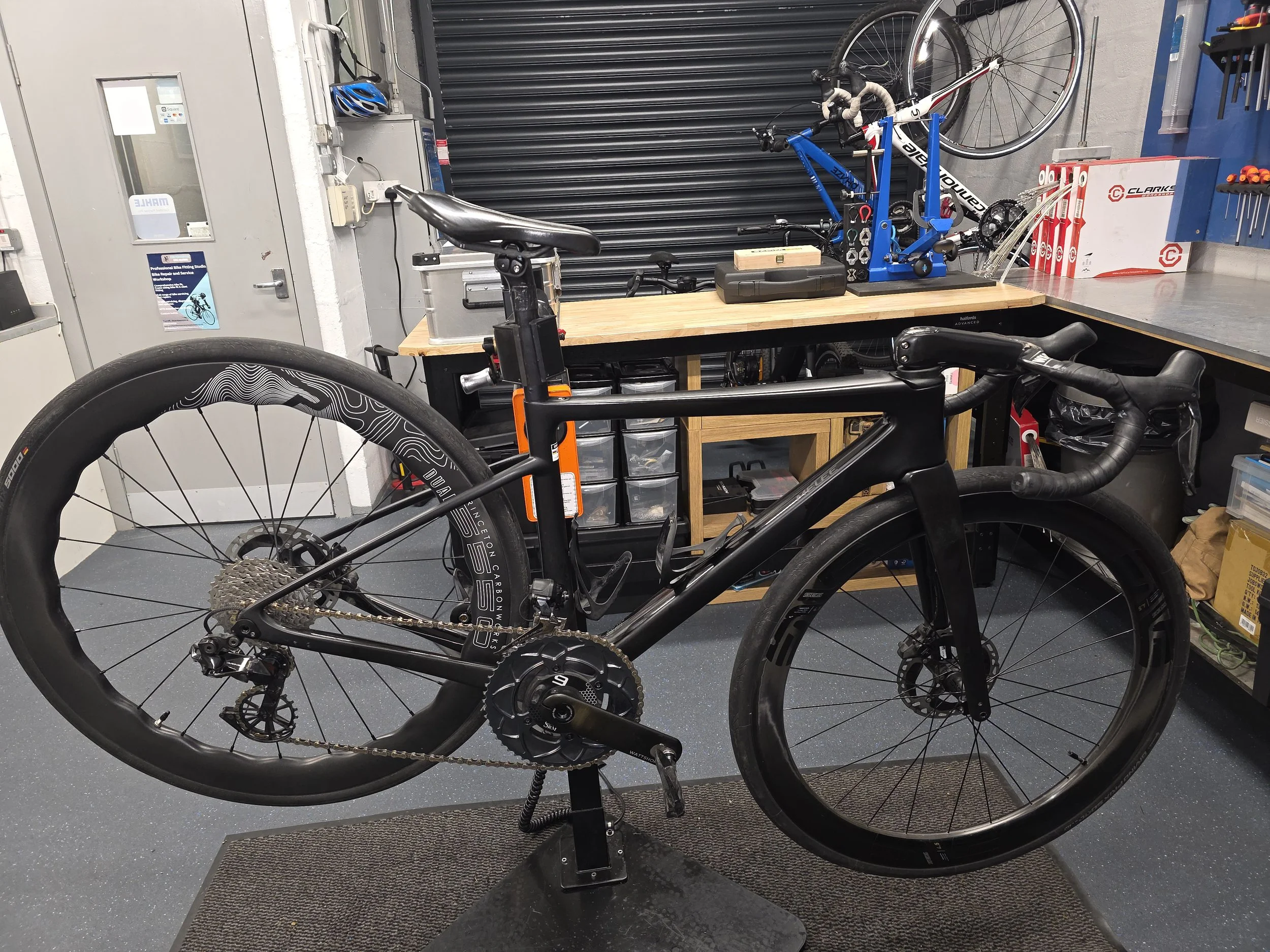 Black road bicycle on a repair stand inside a professional bike fitting and repair workshop, with tools and a second bicycle in the background.