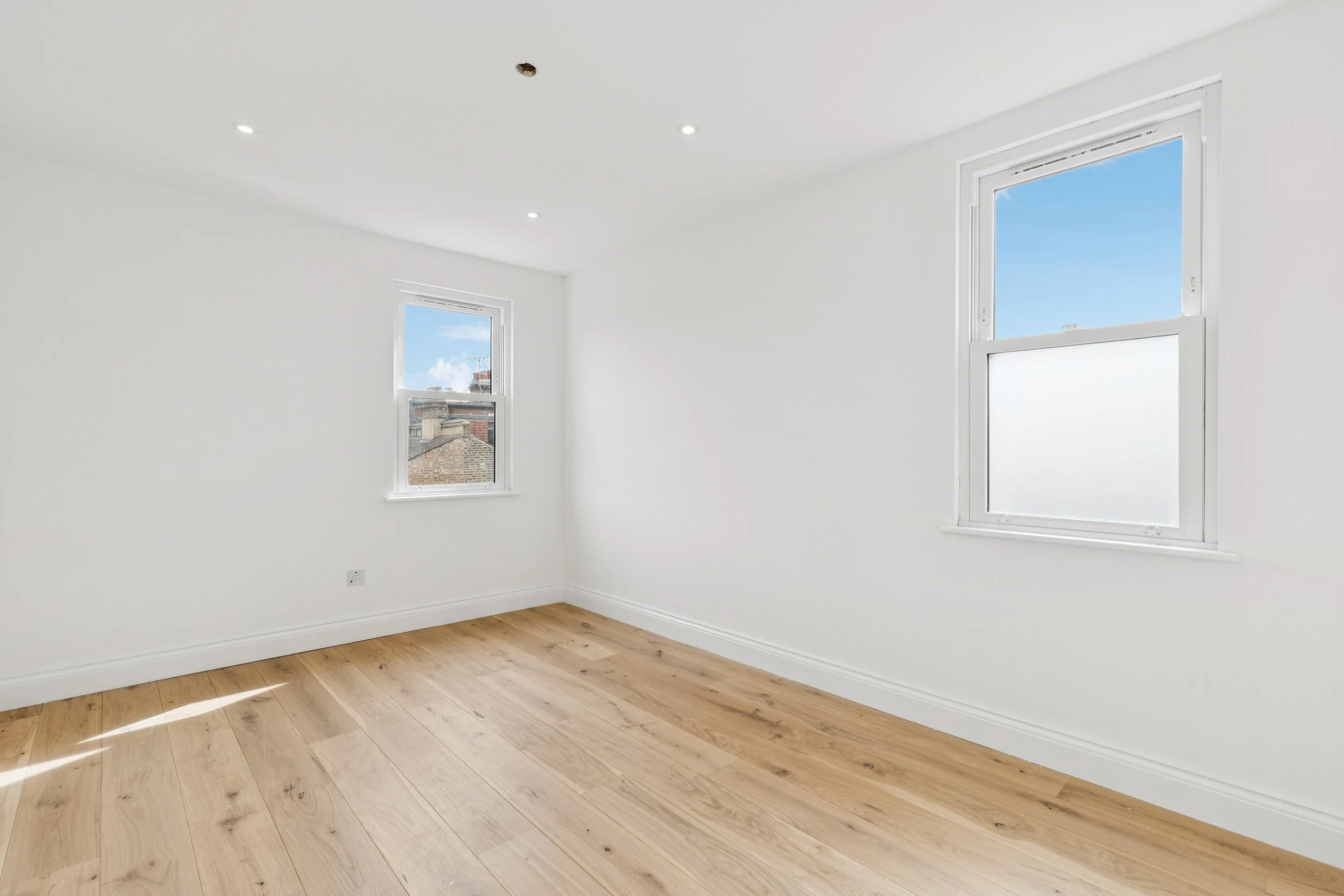 Empty room with white walls, two windows revealing blue sky and city rooftops, and wooden flooring.