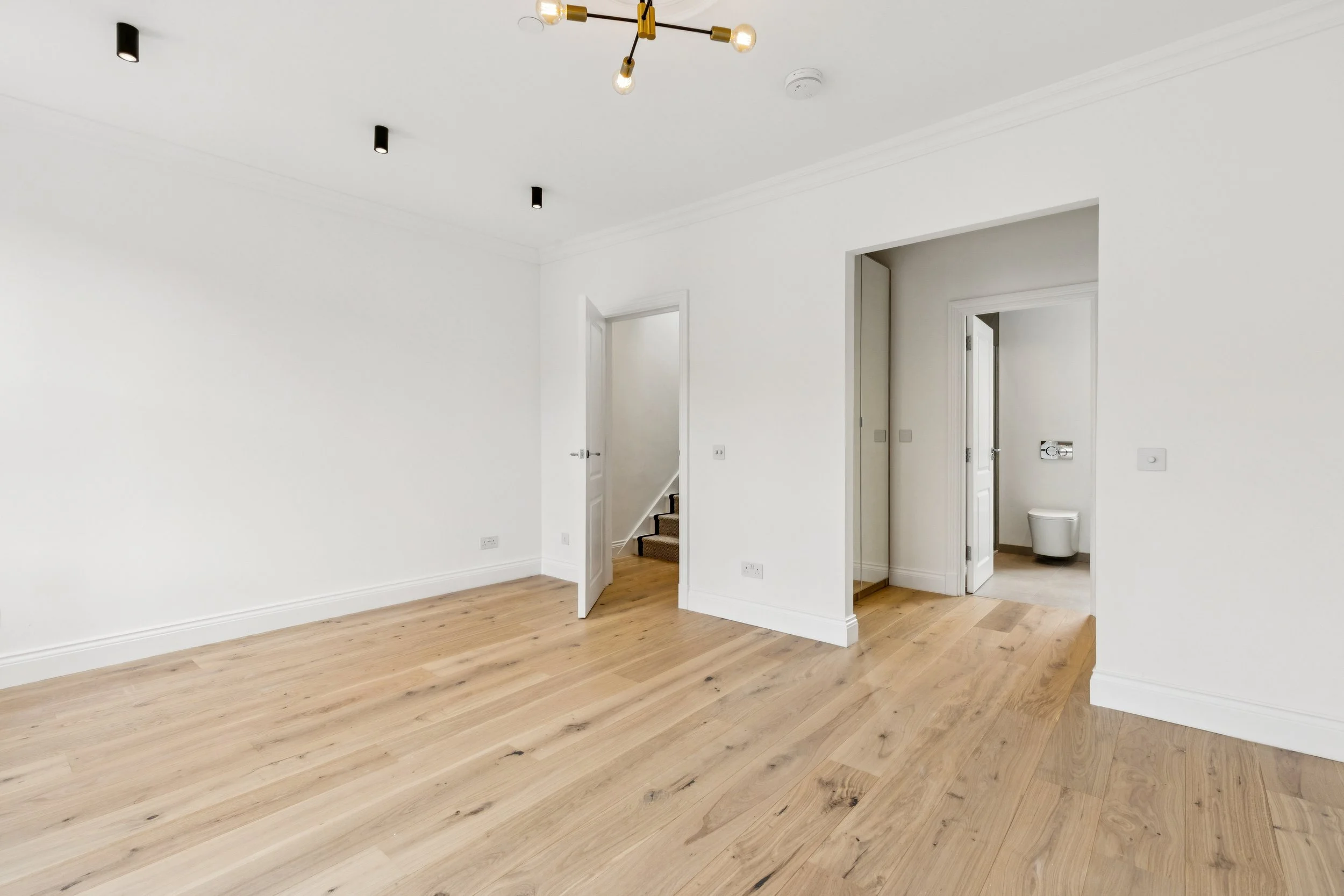Empty room with white walls and wooden flooring, two doors leading to other rooms, one of which shows a toilet.