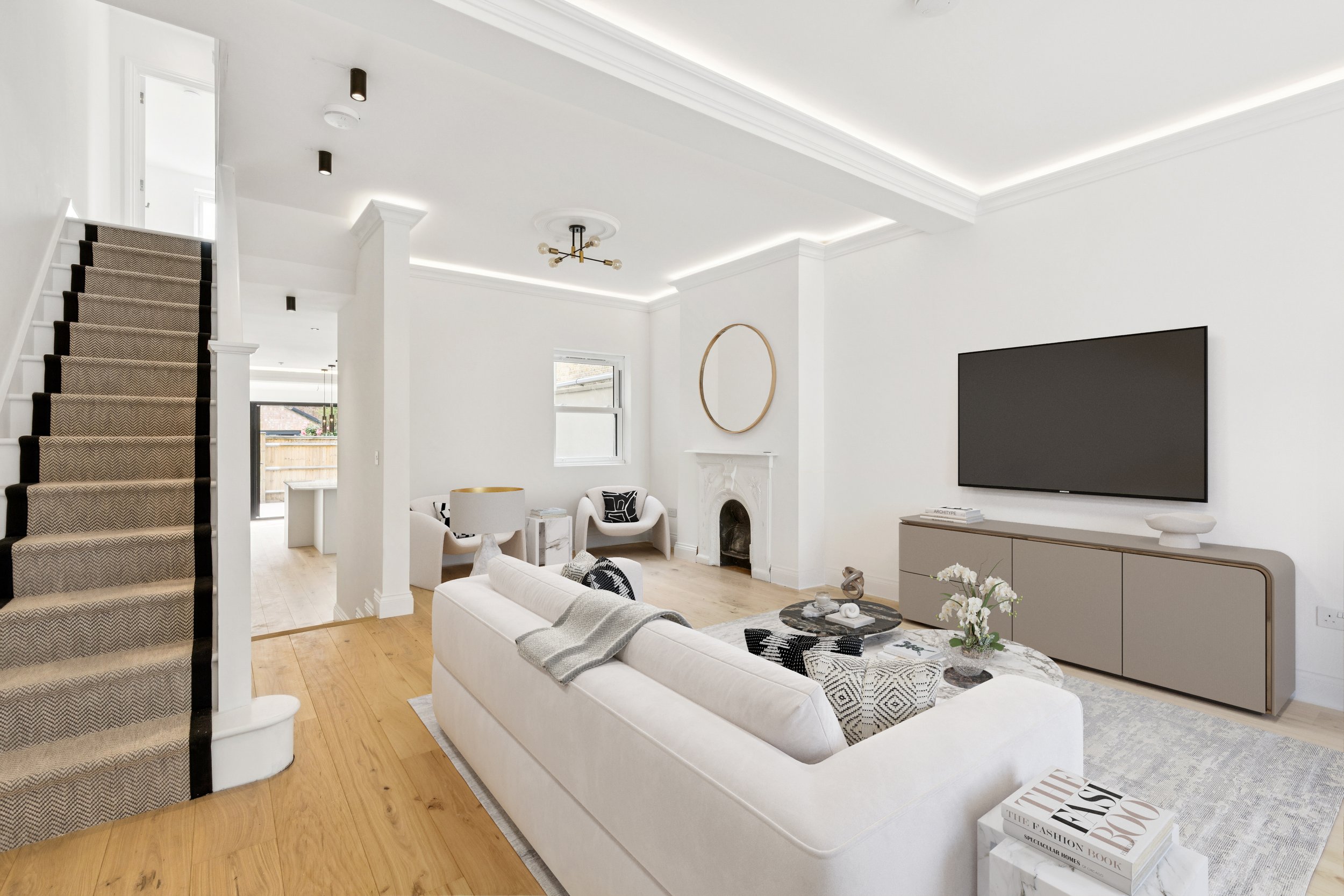 Living room with white walls, hardwood floors, a beige staircase with black accents, modern lighting, and a large flat-screen TV on a beige cabinet. Decor includes a round mirror, chairs, coffee table with books, flowers, and black-and-white cushions