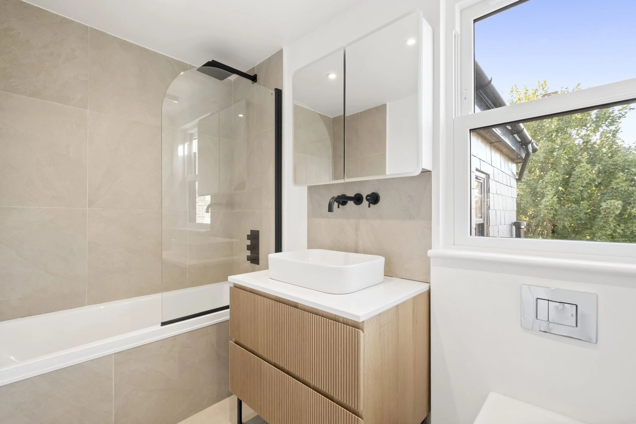 Modern bathroom with beige tiled shower and a black showerhead, a vanity with a white vessel sink, black fixtures, a mirrored cabinet, and a window showing outdoor greenery.
