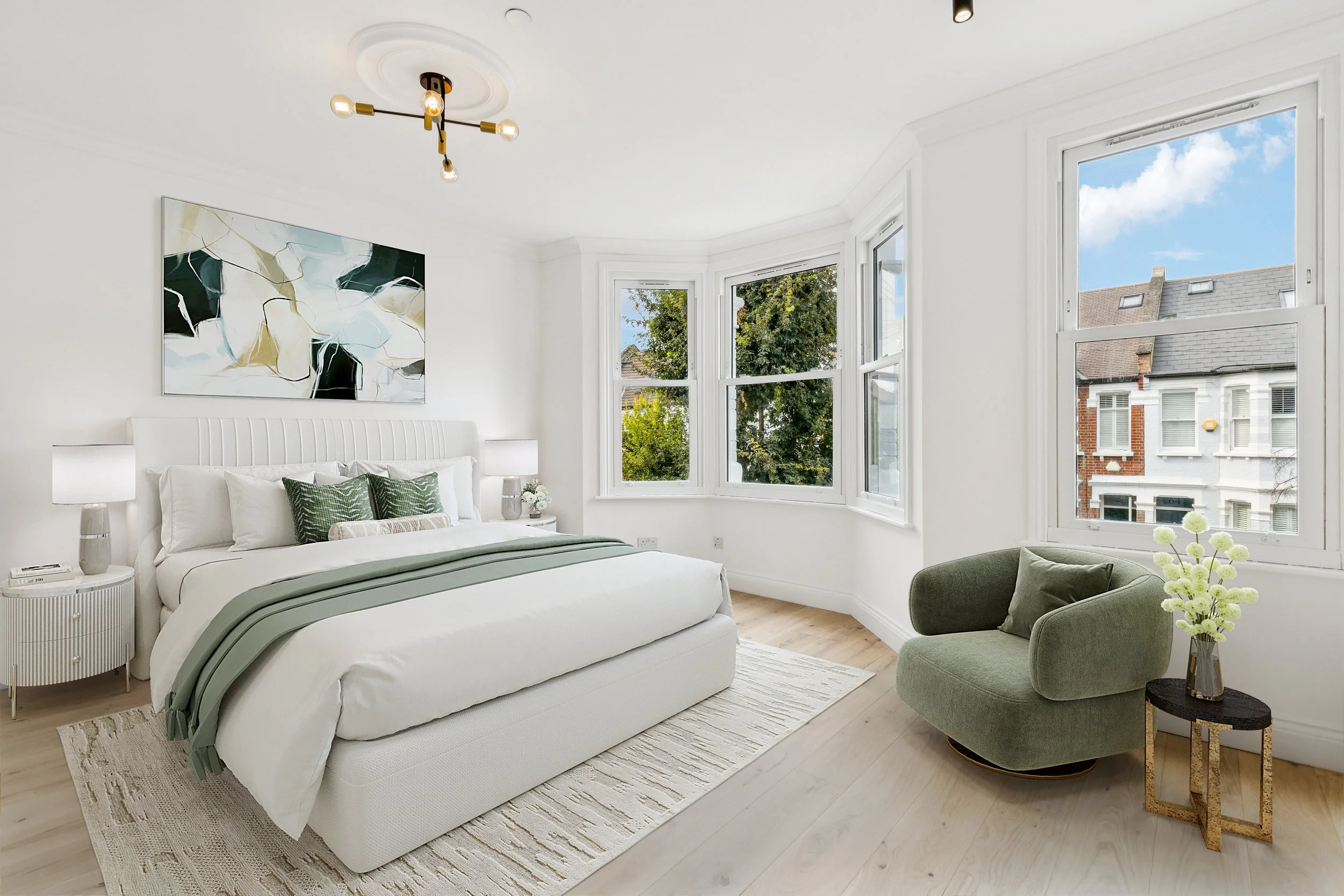 Bright bedroom with white walls and large windows showing a neighborhood. A white bed with green pillows and a gray throw blanket is next to a white nightstand with a lamp. A modern black and gold chandelier hangs from the ceiling. A green armchair a