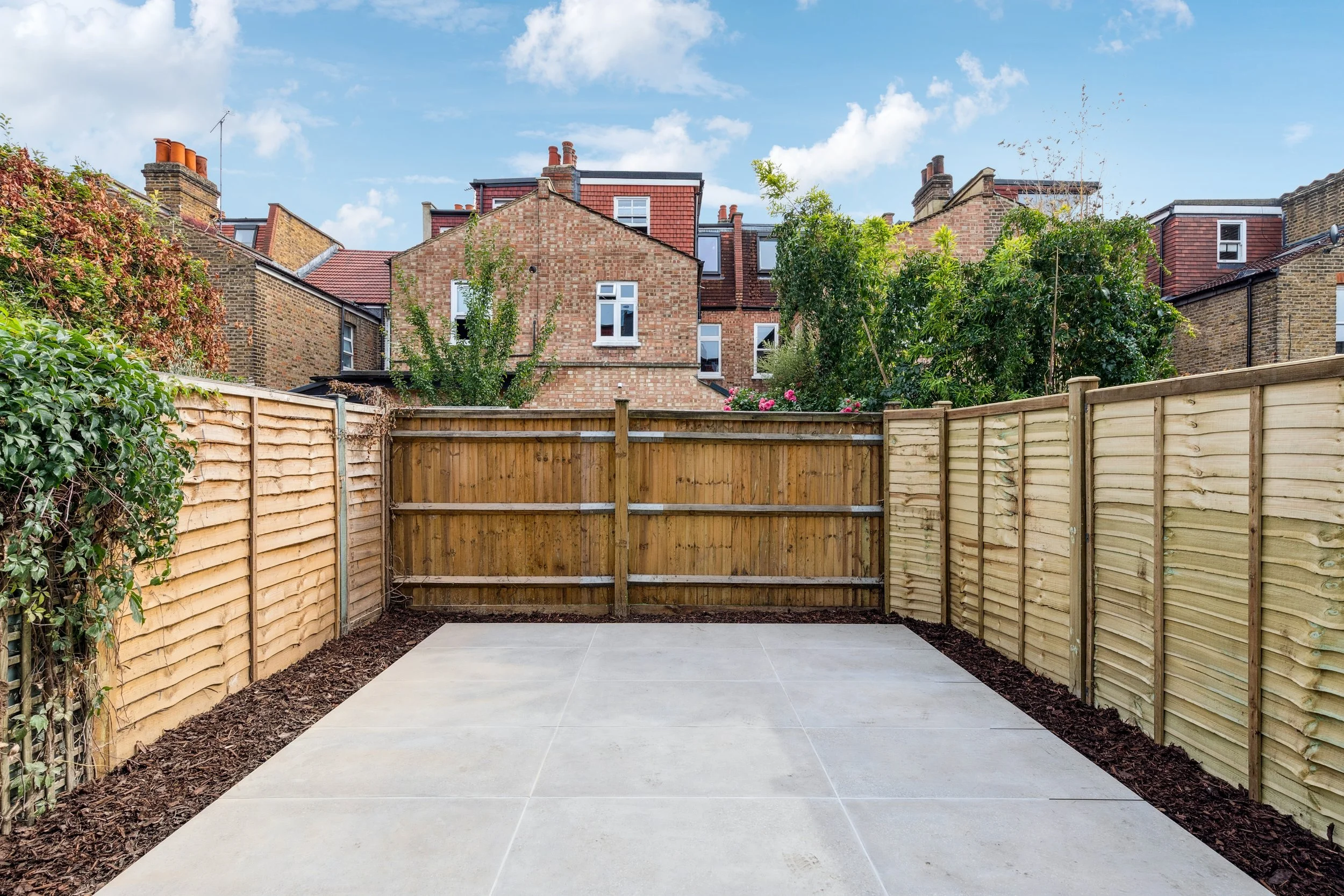 A completed backyard patio with large light gray tiles, surrounded by a new wooden fence, with bushes and trees in the background, under a partly cloudy sky.